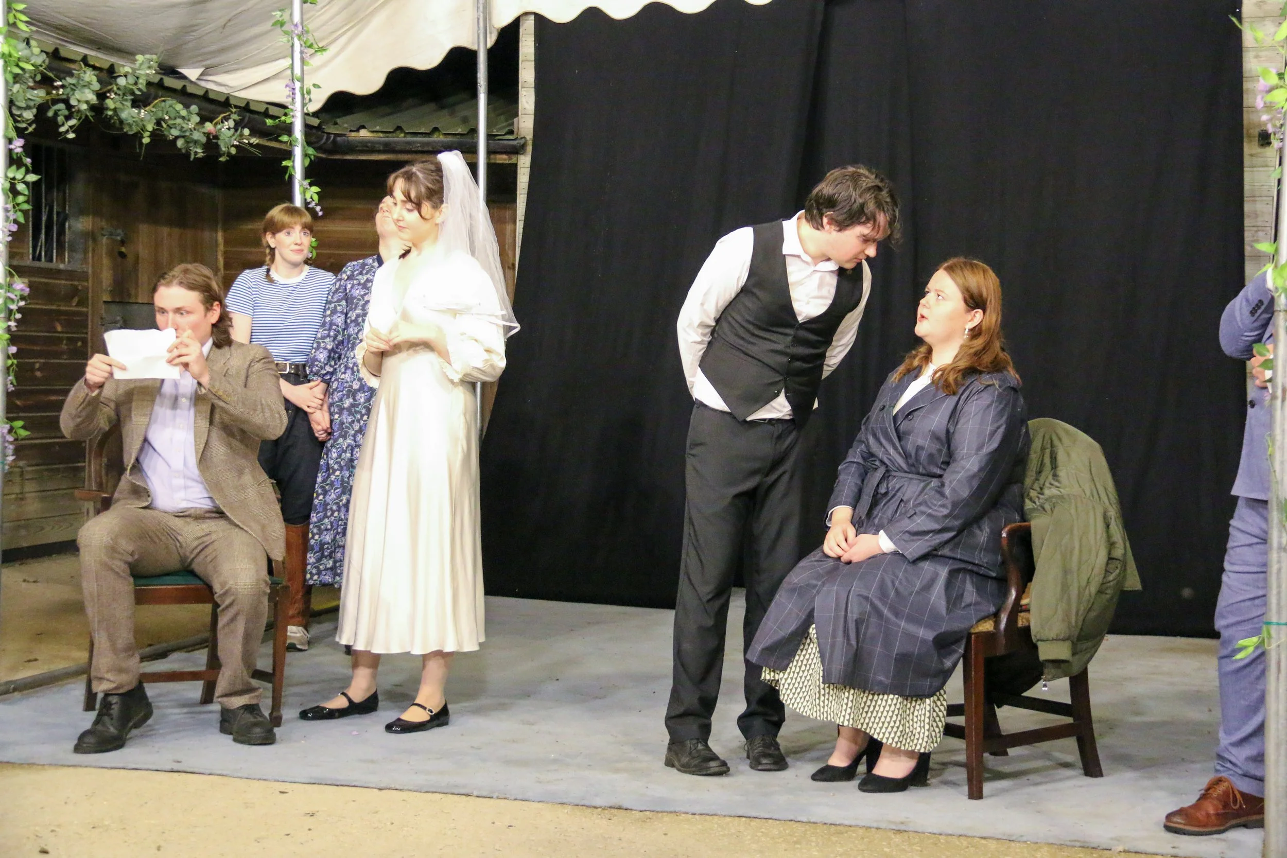 People performing a play on stage, with five performers in period clothing. One woman is seated, another man and woman are standing and talking, and others are engaged in various actions in the background.