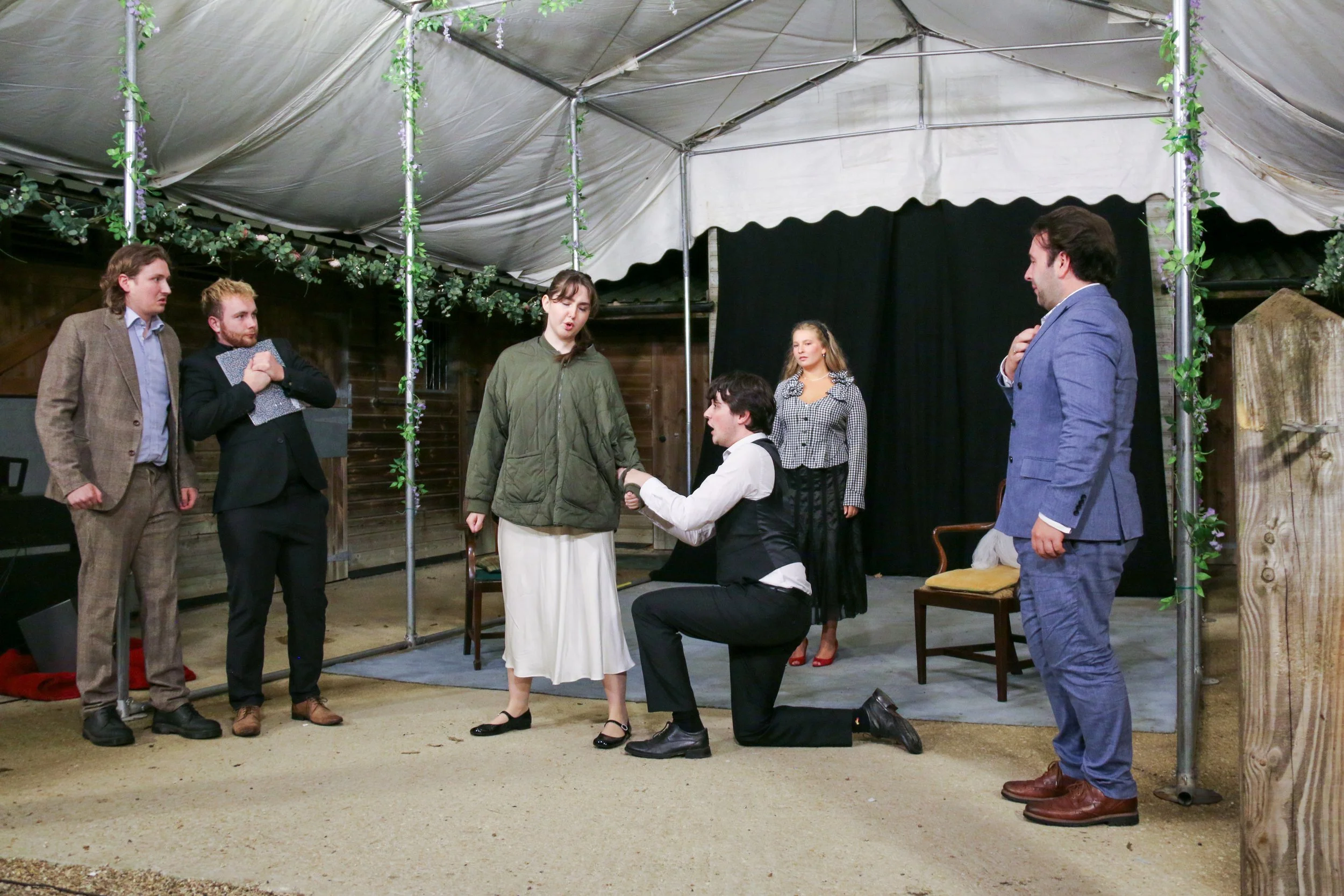 The image shows a theatrical rehearsal with six actors on a stage under a canopy, with one actor kneeling and holding the hand of a female actor standing before him, while four other actors watch. The setting appears to be an indoor or semi-outdoor s