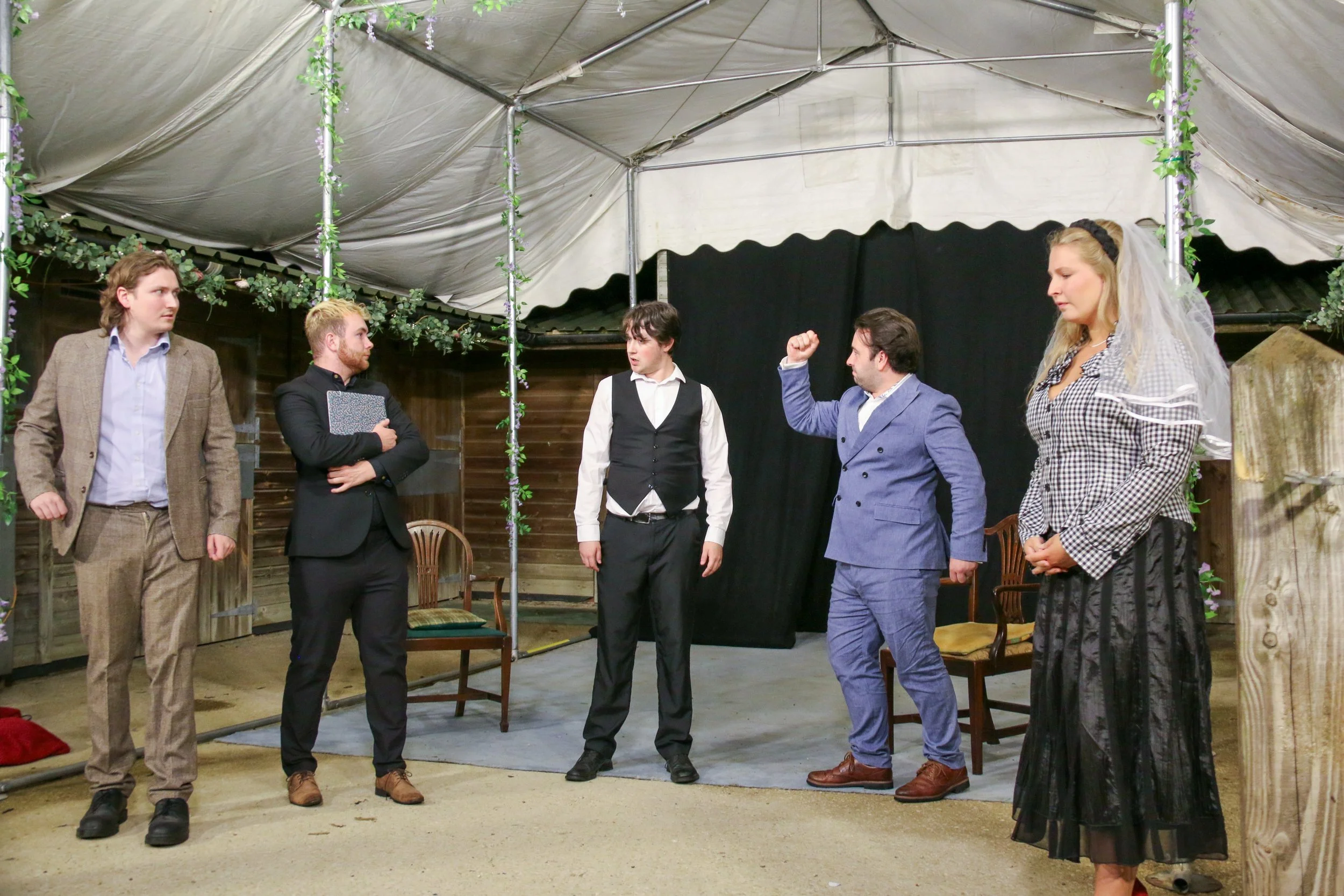 Six people dressed in formal and semi-formal attire standing on a stage with a black curtain backdrop, inside a tent decorated with artificial flowers and greenery.