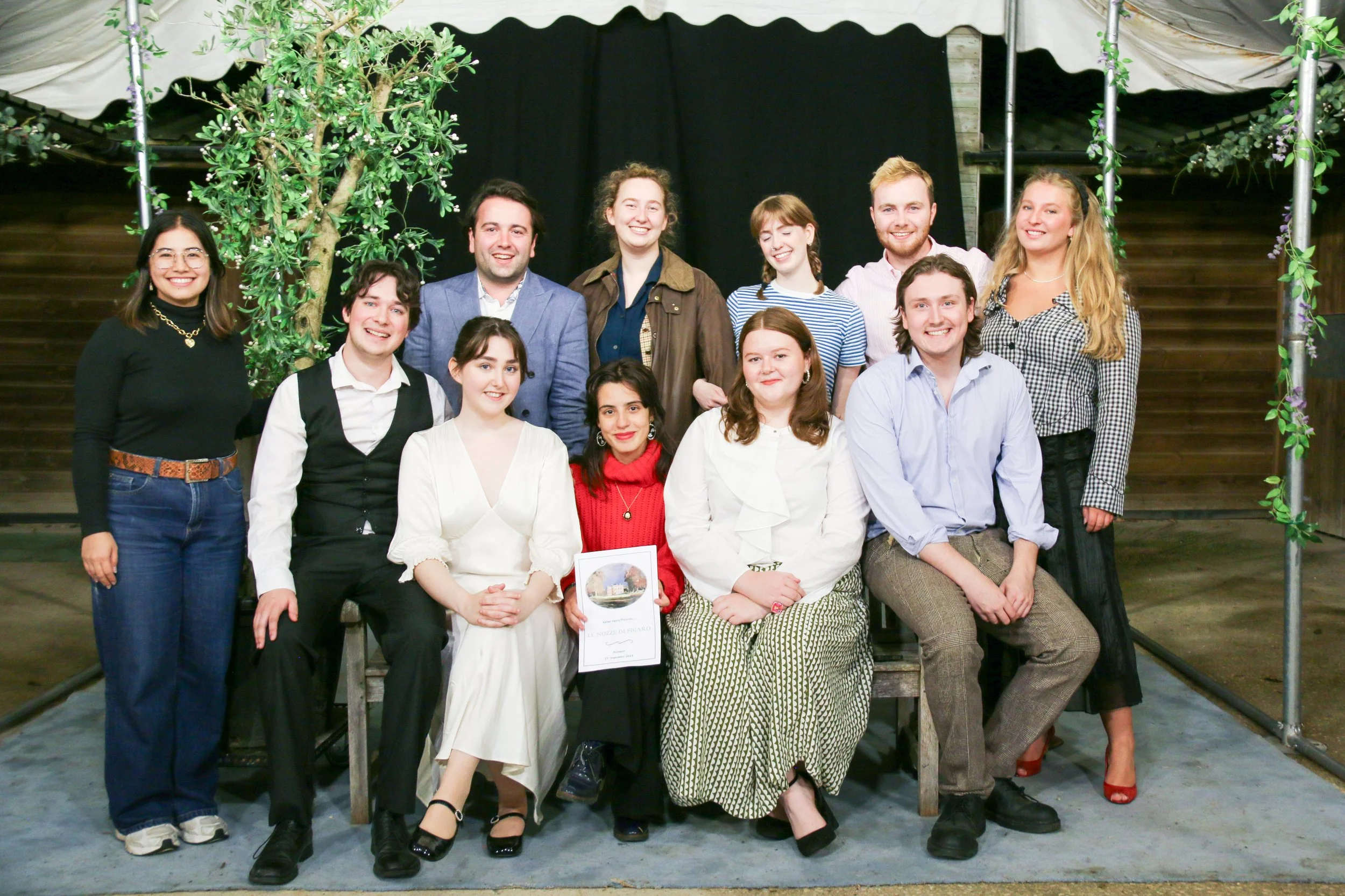 A group of fifteen young adults posing for a photo on a stage with a black backdrop, some standing and some seated, in an indoor setting.