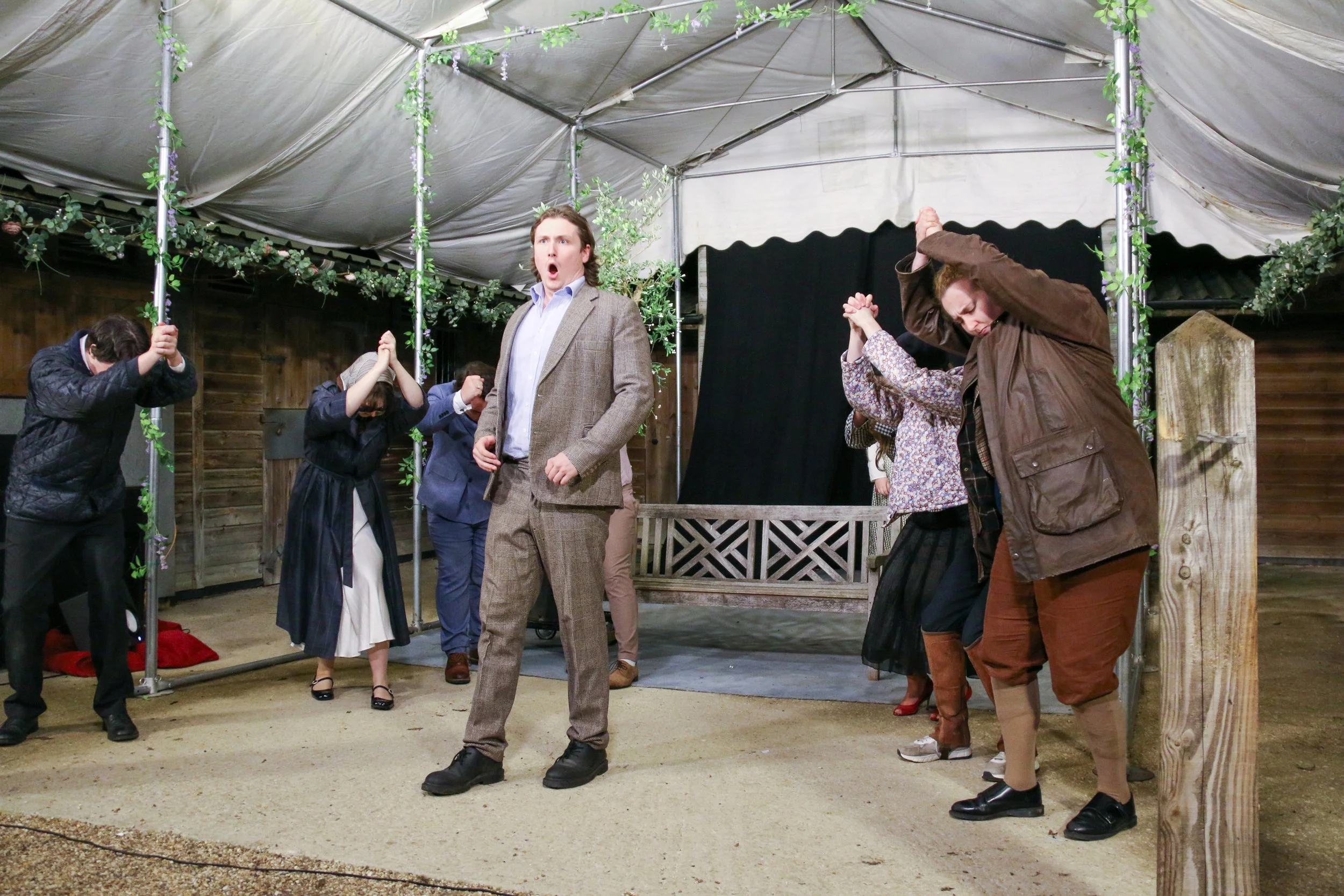 A group of people are standing inside a decorated tent, appearing to be praying or participating in a ritual. The scene is lively, with various individuals engaging with their hands raised or clasped, and a man in a suit in the center looking surpris