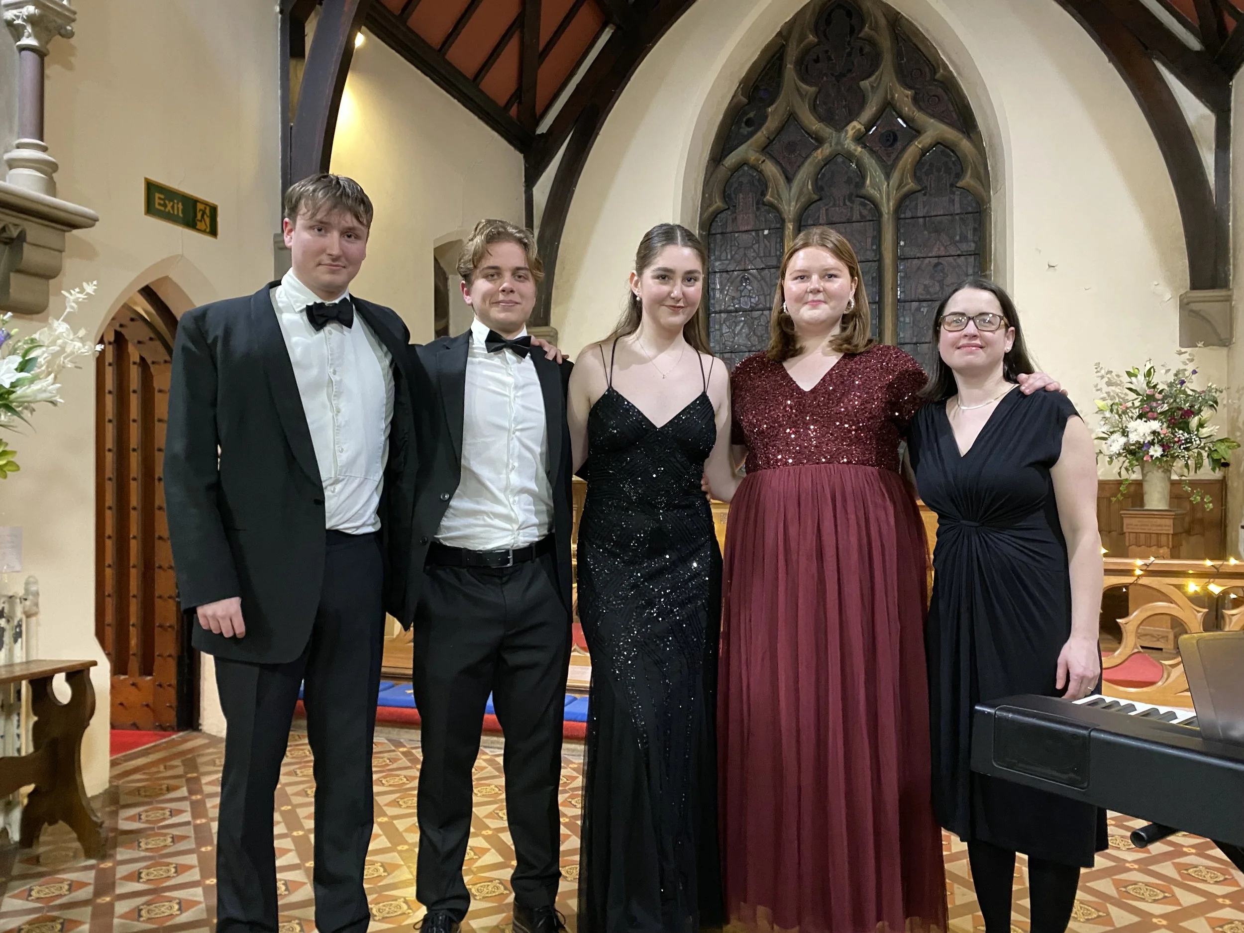 Five people dressed in formal attire standing inside a church with stained glass windows, flowers, and a keyboard in the foreground.