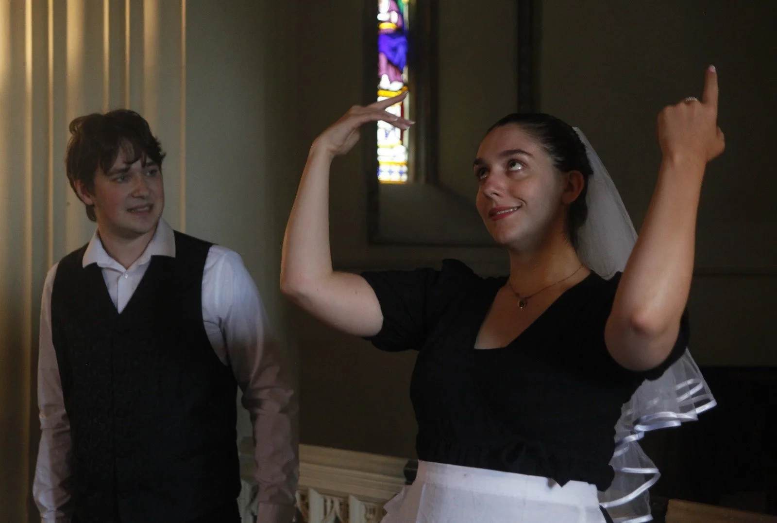 A woman dressed as a bride making a gesture with her hands while a young man dressed in a vest and shirt looks at her inside a church with stained glass window.