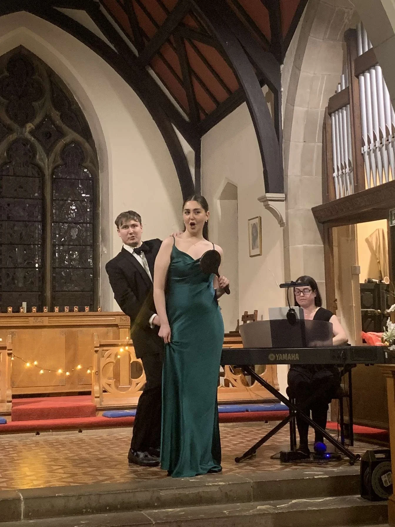 A young woman in a green gown holding a small mirror sings on stage, accompanied by a man in a tuxedo standing behind her, in a church or chapel with stained glass windows and an organ, while a pianist plays on a Yamaha keyboard.