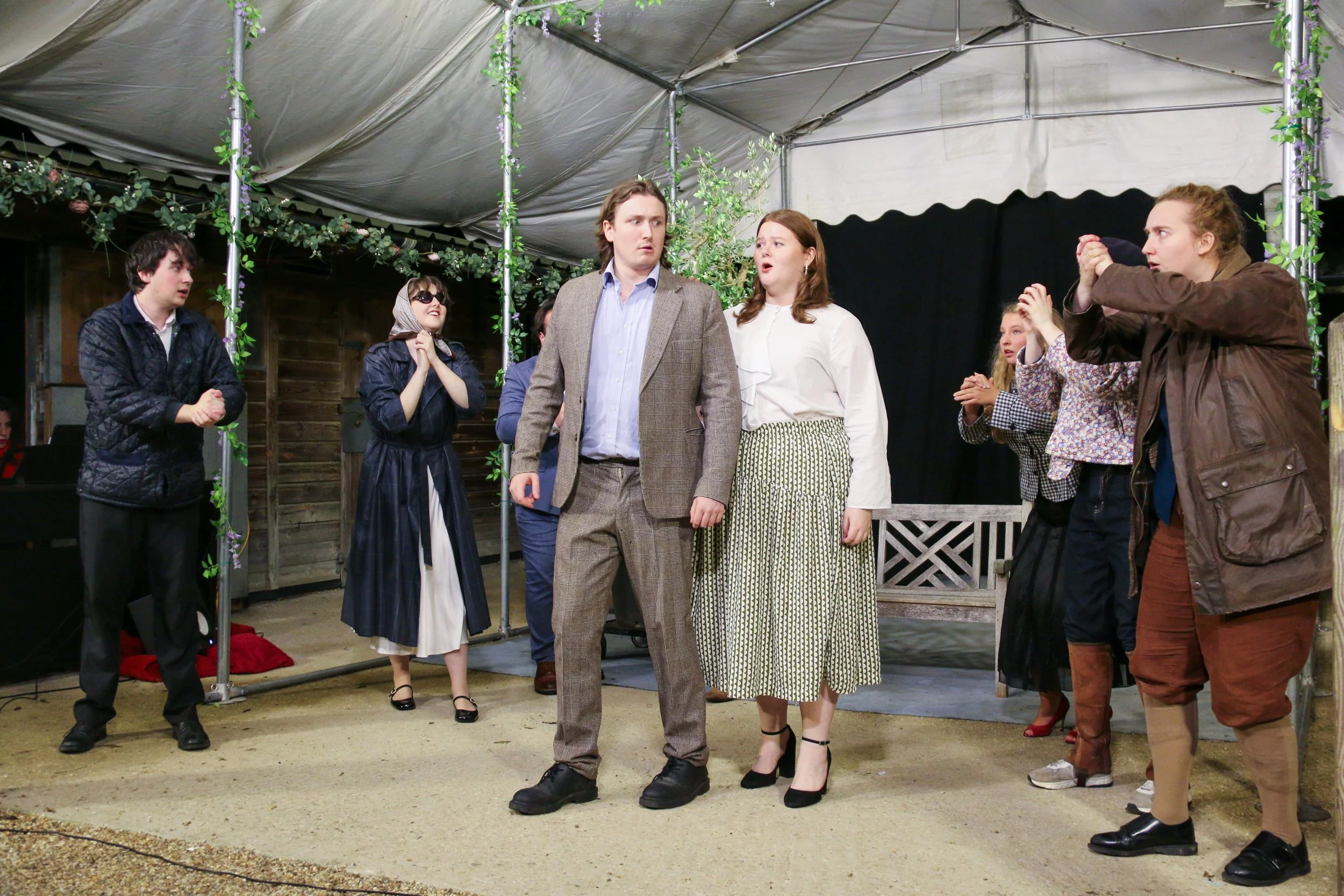 A group of actors on a stage performing a play, with some in costume and others in casual clothing, under a tent with decorative greenery.