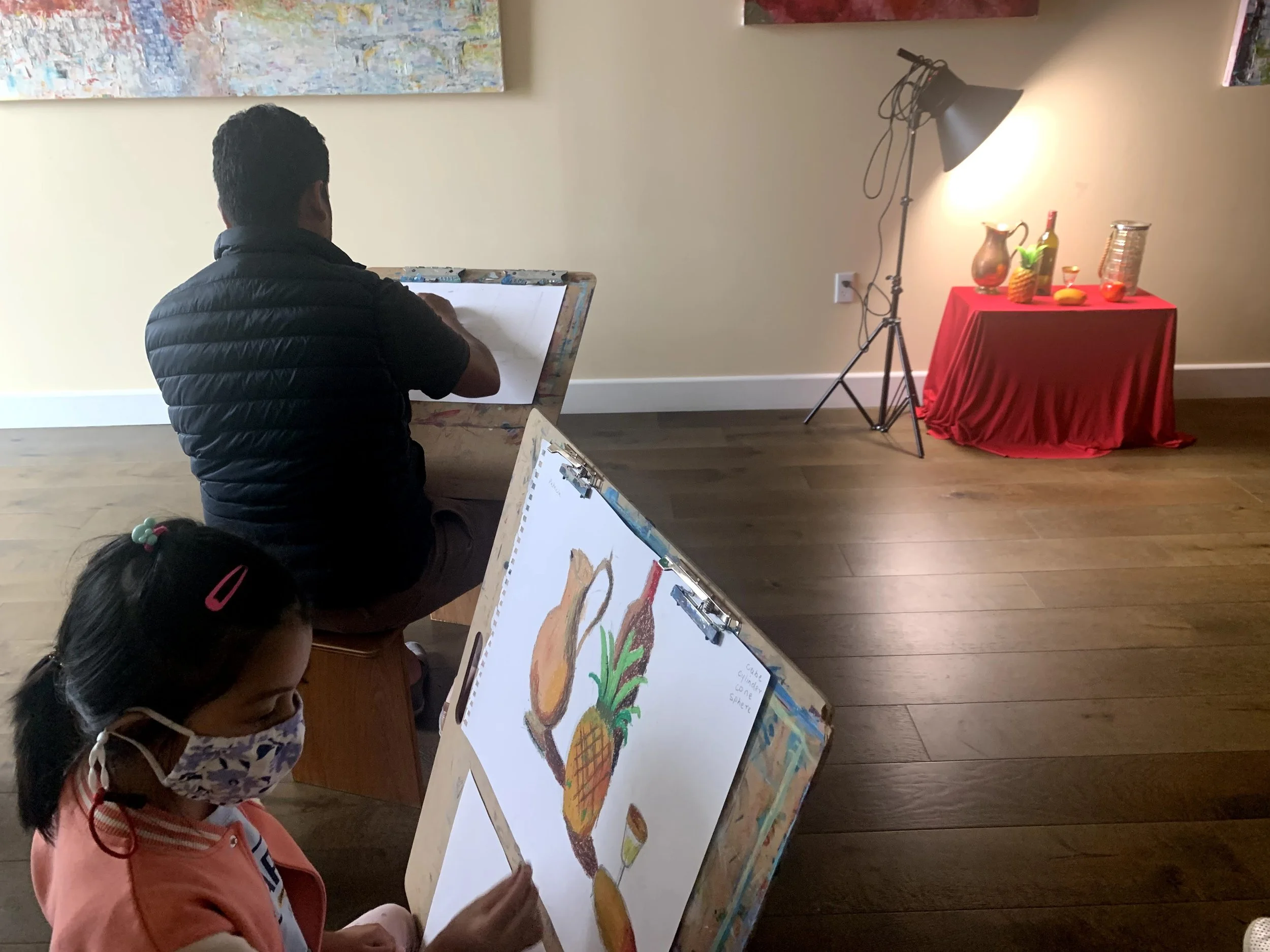 A woman and a man painting on canvases in an art studio. The woman is sitting in front, wearing a pink shirt and a face mask, holding a paintbrush. The man is sitting beside her, painting on a vertical canvas, wearing a black vest. Behind them is a w