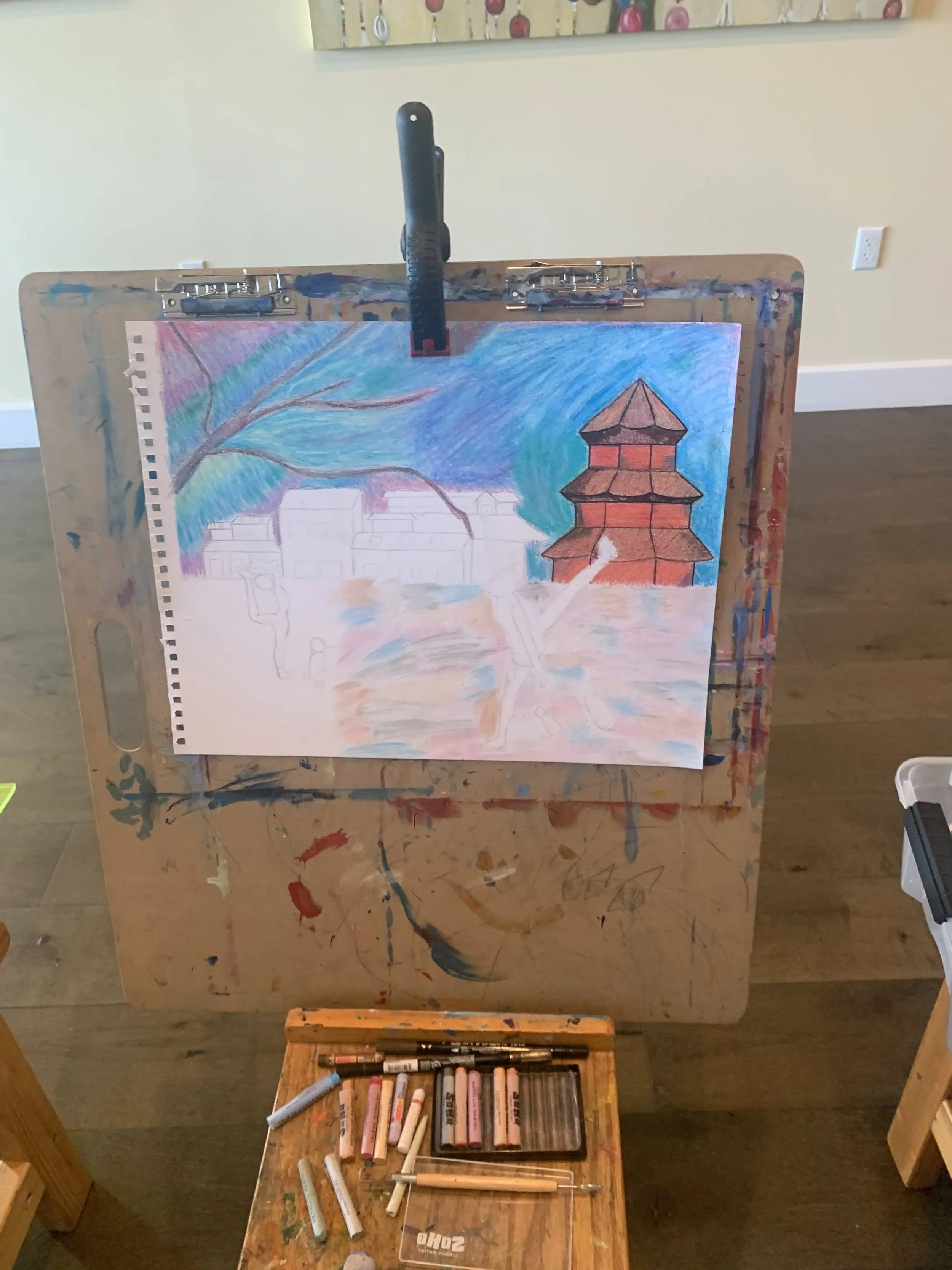 A partially completed colorful landscape painting on a wooden easel, featuring a tree with sprawling branches, a red multi-tiered pagoda-style building, and a blue sky, with art supplies on a small wooden table in front.