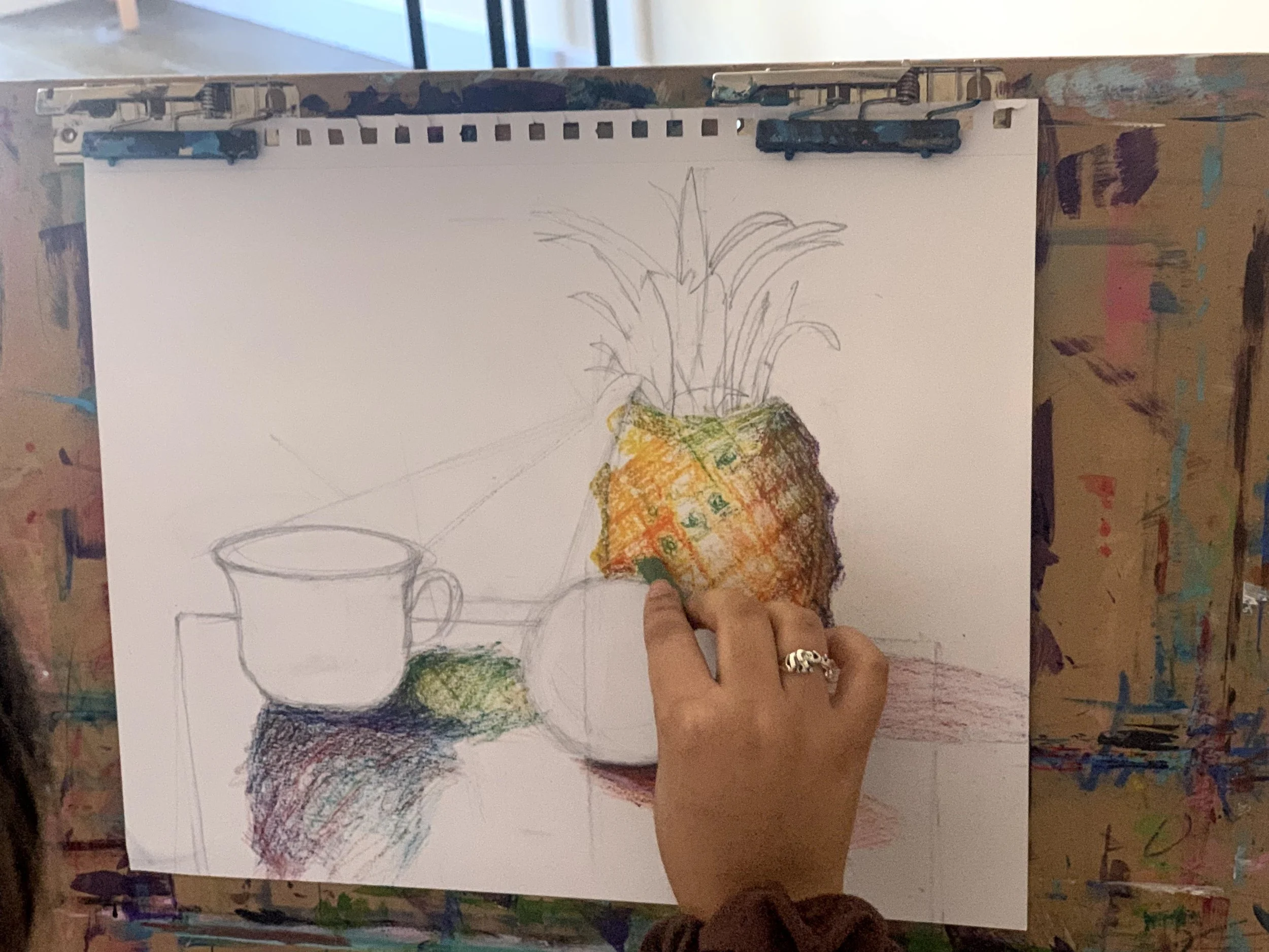 A person is sketching a still life of a pineapple, a cup, and a bowl on a white sheet of paper. The person is holding a crayon over the pineapple in the drawing.