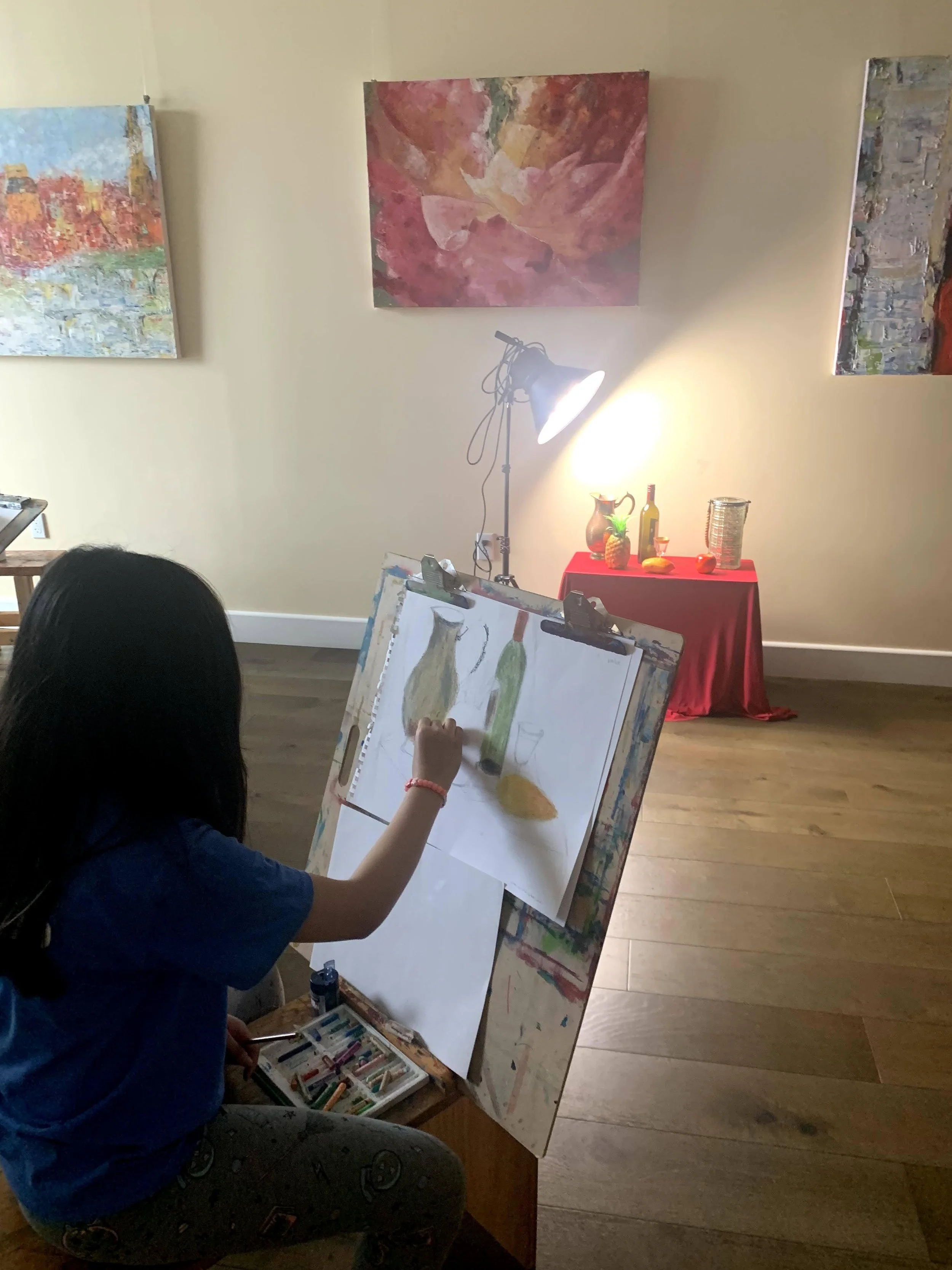 A young girl drawing still life objects on an easel in an art studio, with paintings on the wall and a red table with bottles and decorative items in the background.