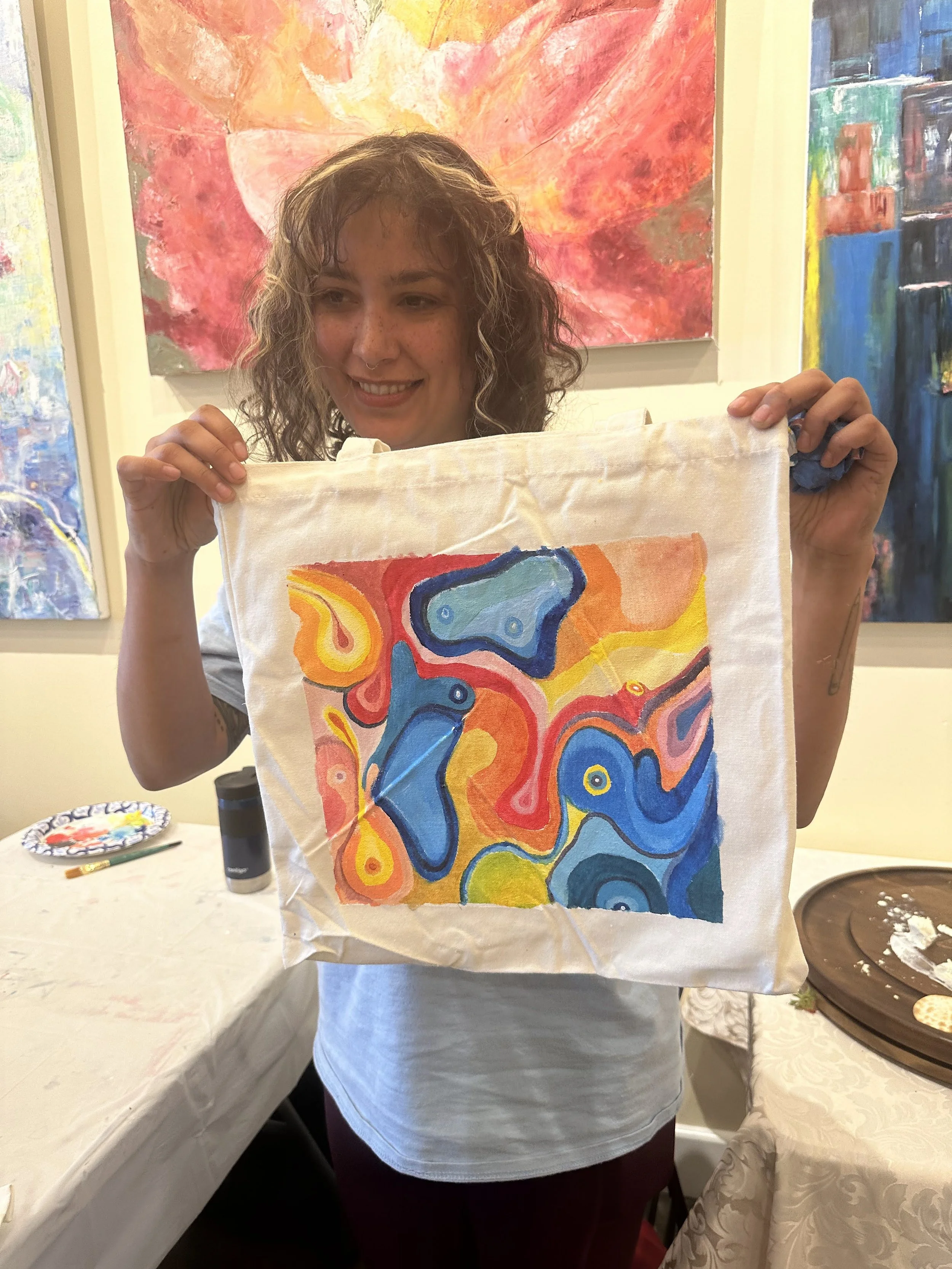 A woman with curly hair smiling and holding up a colorful abstract painting on fabric in an art gallery.
