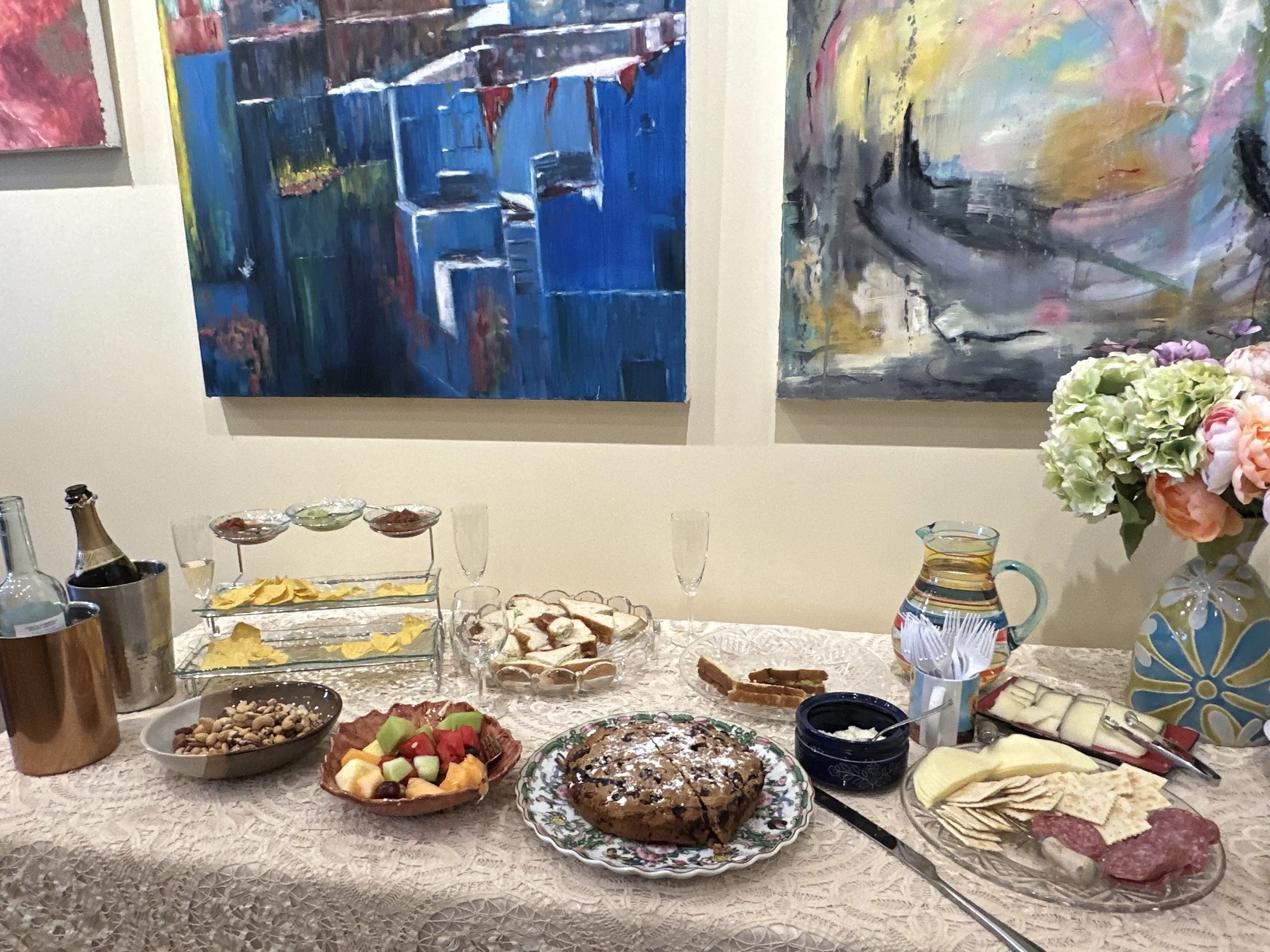A table set with various snacks and drinks, including a fruit platter, a cake, cheese, crackers, nuts, and beverages, with colorful abstract paintings on the wall behind.
