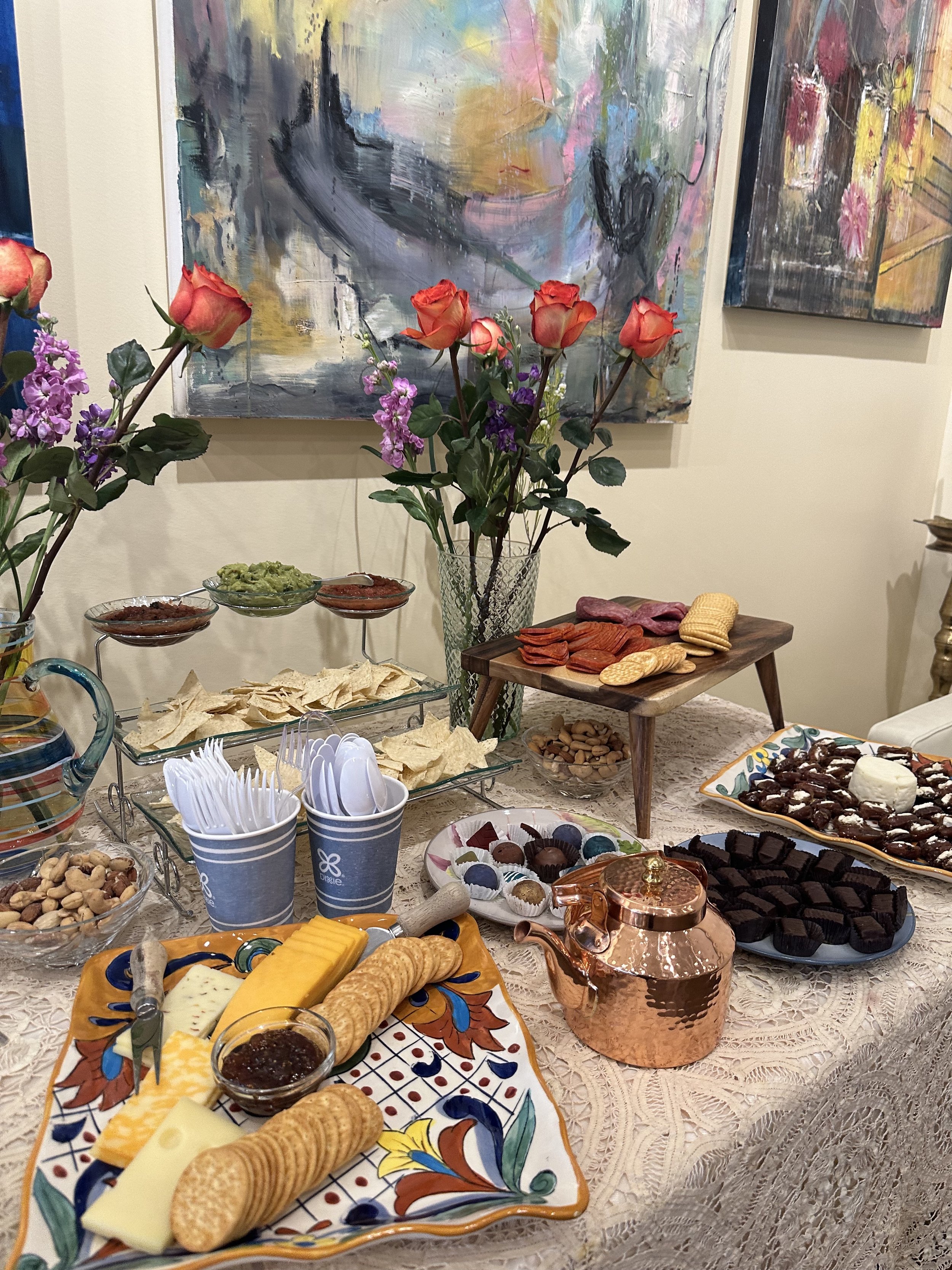 A table set with assorted cheeses, crackers, chocolates, and dips, featuring a centerpiece of roses and purple flowers, with abstract paintings in the background.