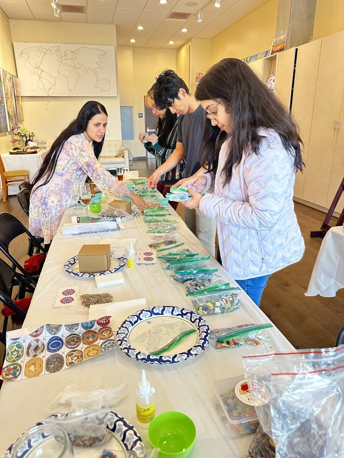 People standing around a table, assembling colorful mosaic art pieces with various tools and supplies in a bright room.