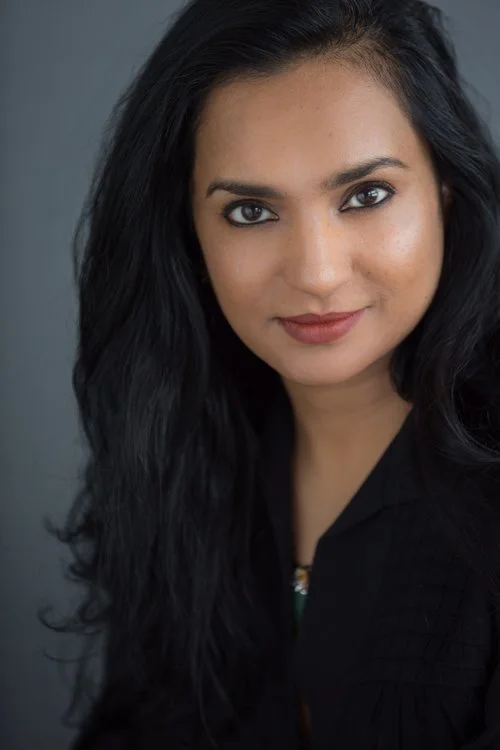 Portrait of a woman with long black hair and a slight smile, wearing a black top, against a gray background.