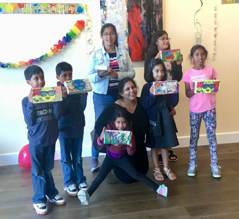 Group of children and a woman at a birthday celebration, holding decorated boxes, with birthday decorations and a rainbow-colored paper garland on the wall.