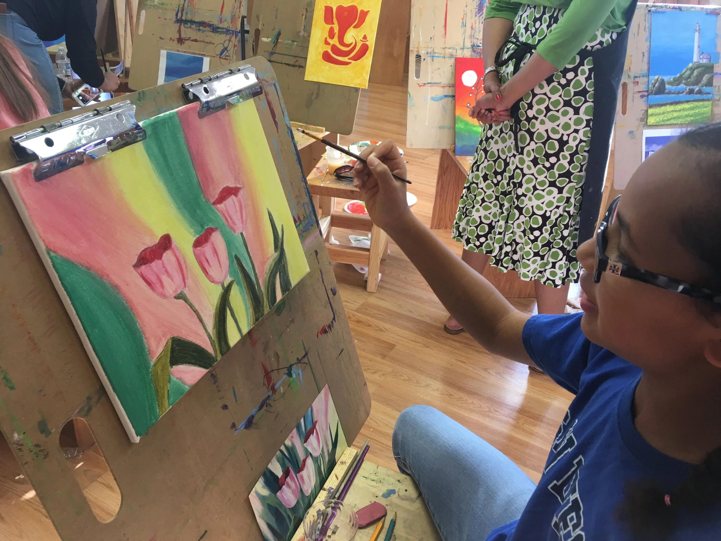 A person wearing glasses and a blue shirt is painting pink tulips on a canvas set on an easel, in an art studio with colorful paintings and a wooden floor.