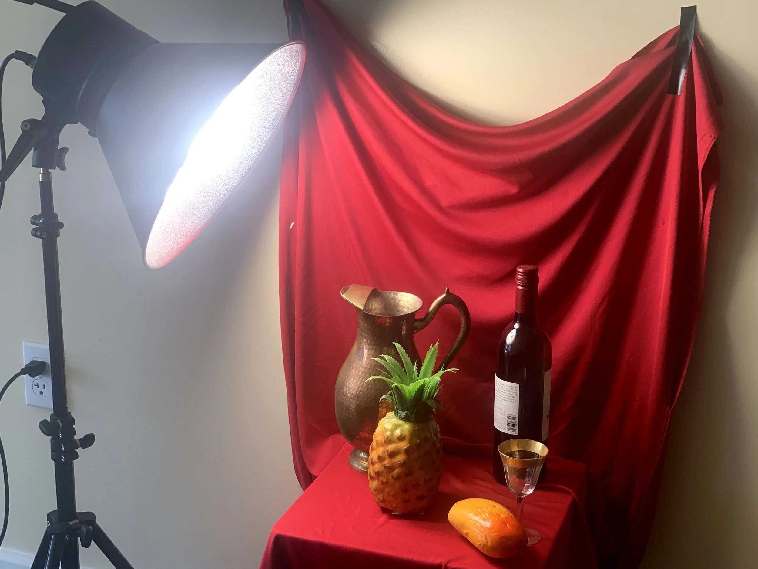 Table with a decorative metal pitcher, a pineapple-shaped container, a bottle of red wine, a wine glass, a mango, and a small potted plant, with a red draped fabric backdrop and studio lighting.