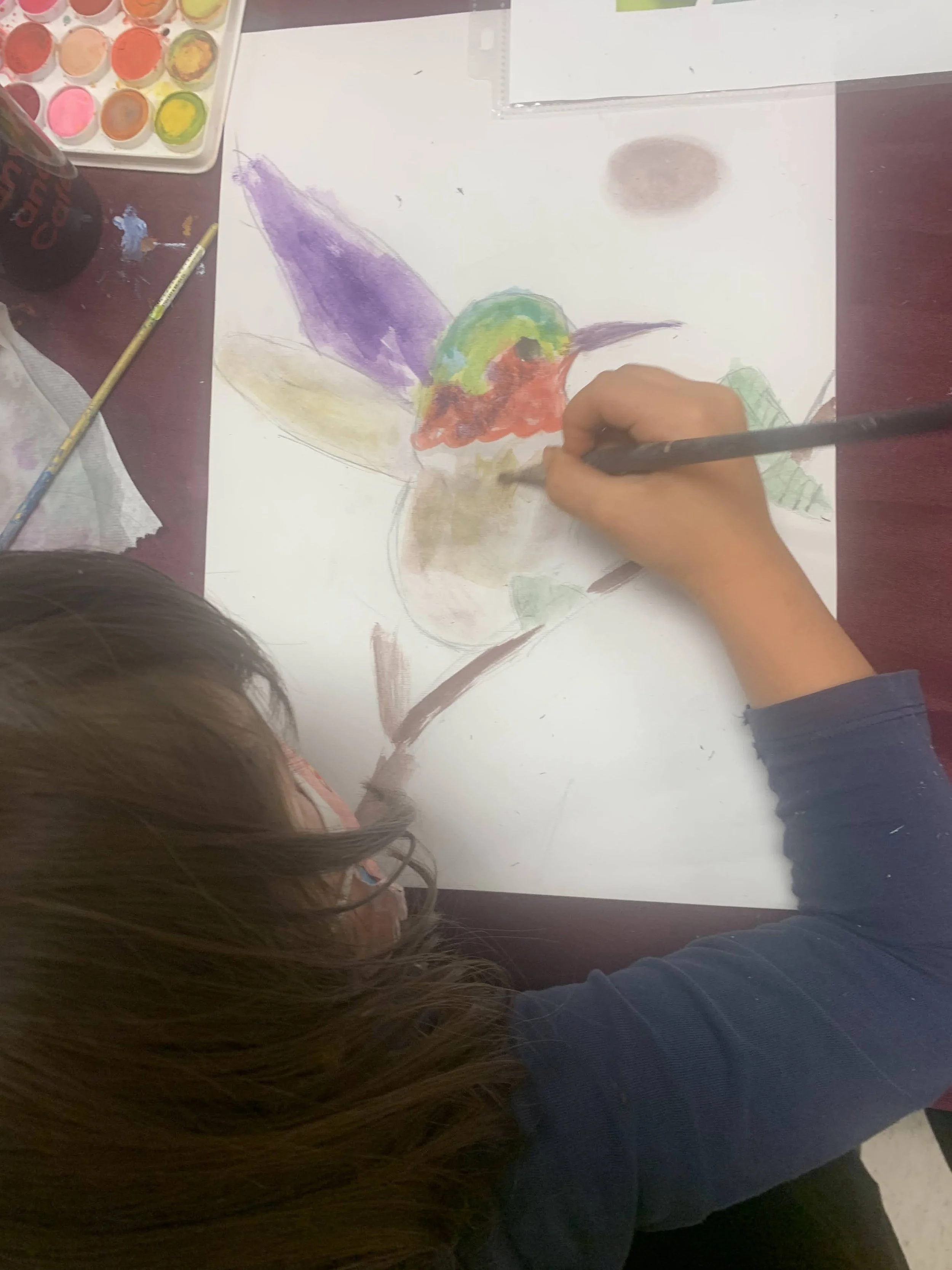 A child is painting a colorful hummingbird on a piece of paper, surrounded by painting supplies.