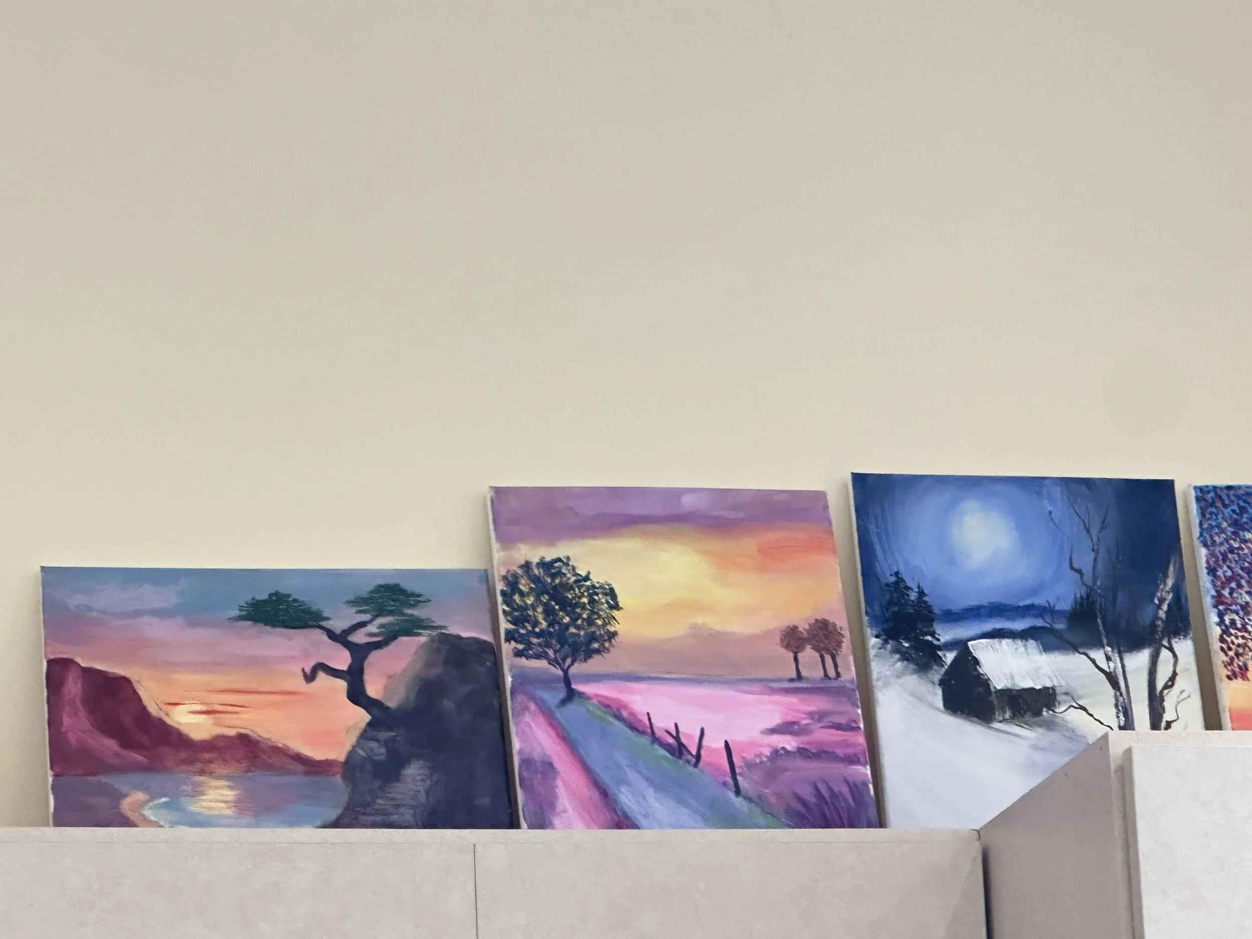 Four landscape paintings leaning against a beige wall. The first depicts a sunset over a river and cliffs with a tree, the second shows a sunset with purple and pink fields and a tree-lined path, the third features a moonlit night scene with a house 