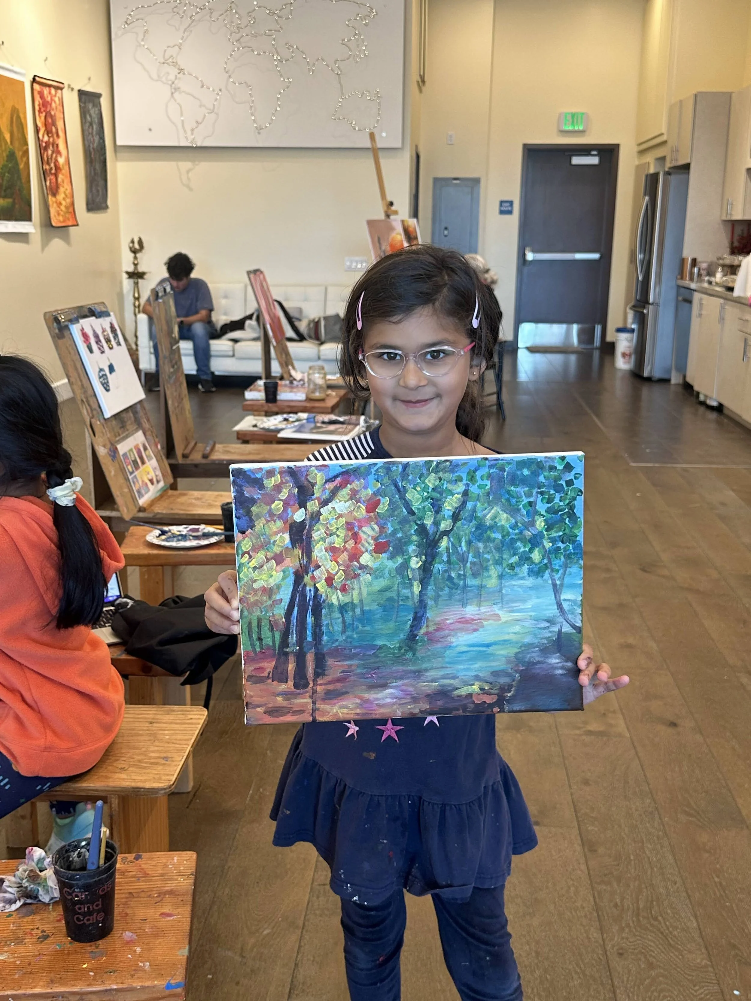Young girl in glasses holding a colorful landscape painting of trees and a body of water in an art studio.
