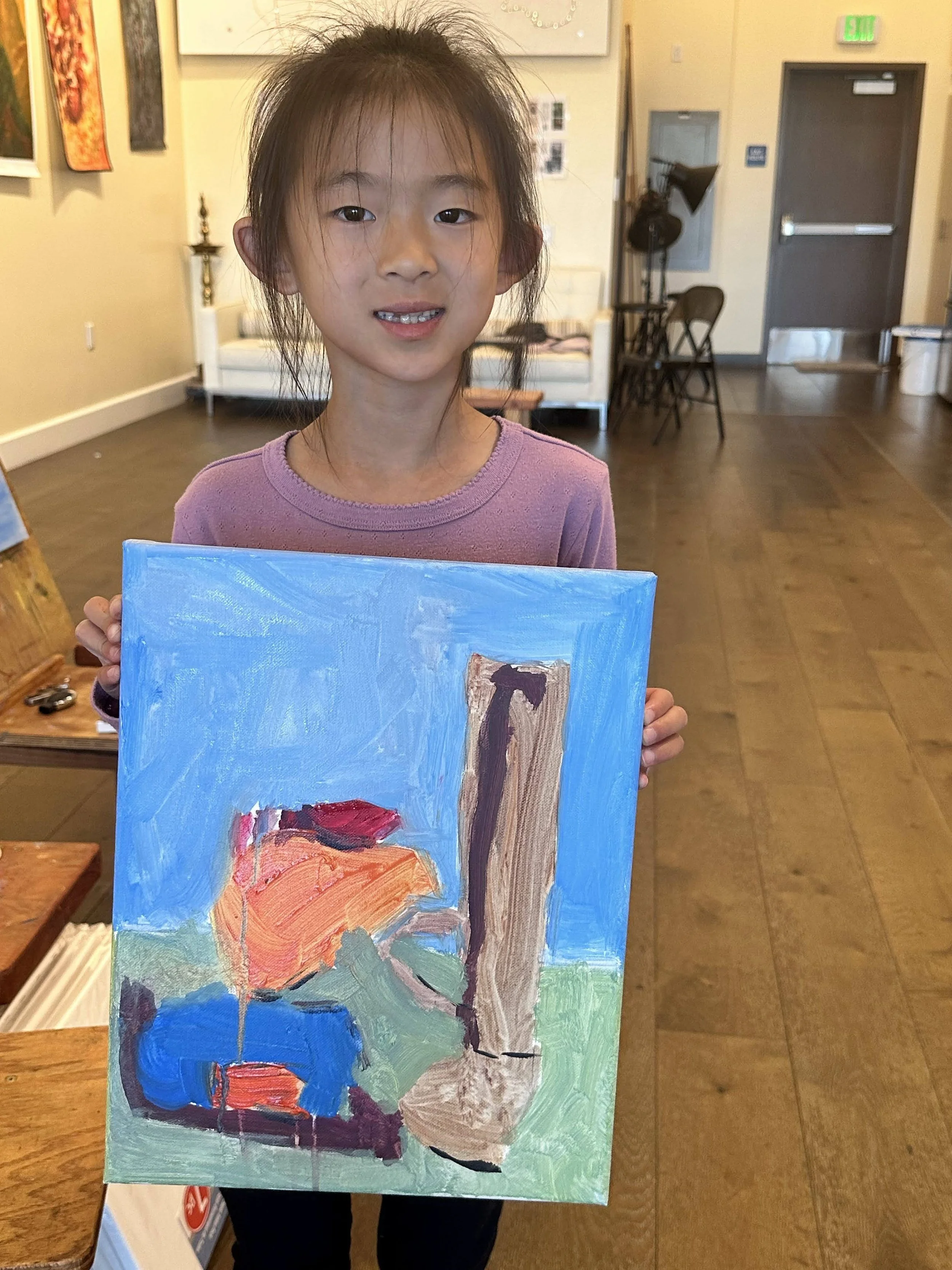 A young girl with messy hair holding up an abstract painting with a blue background, an orange shape, and other colorful brushstrokes in an art gallery or classroom setting.