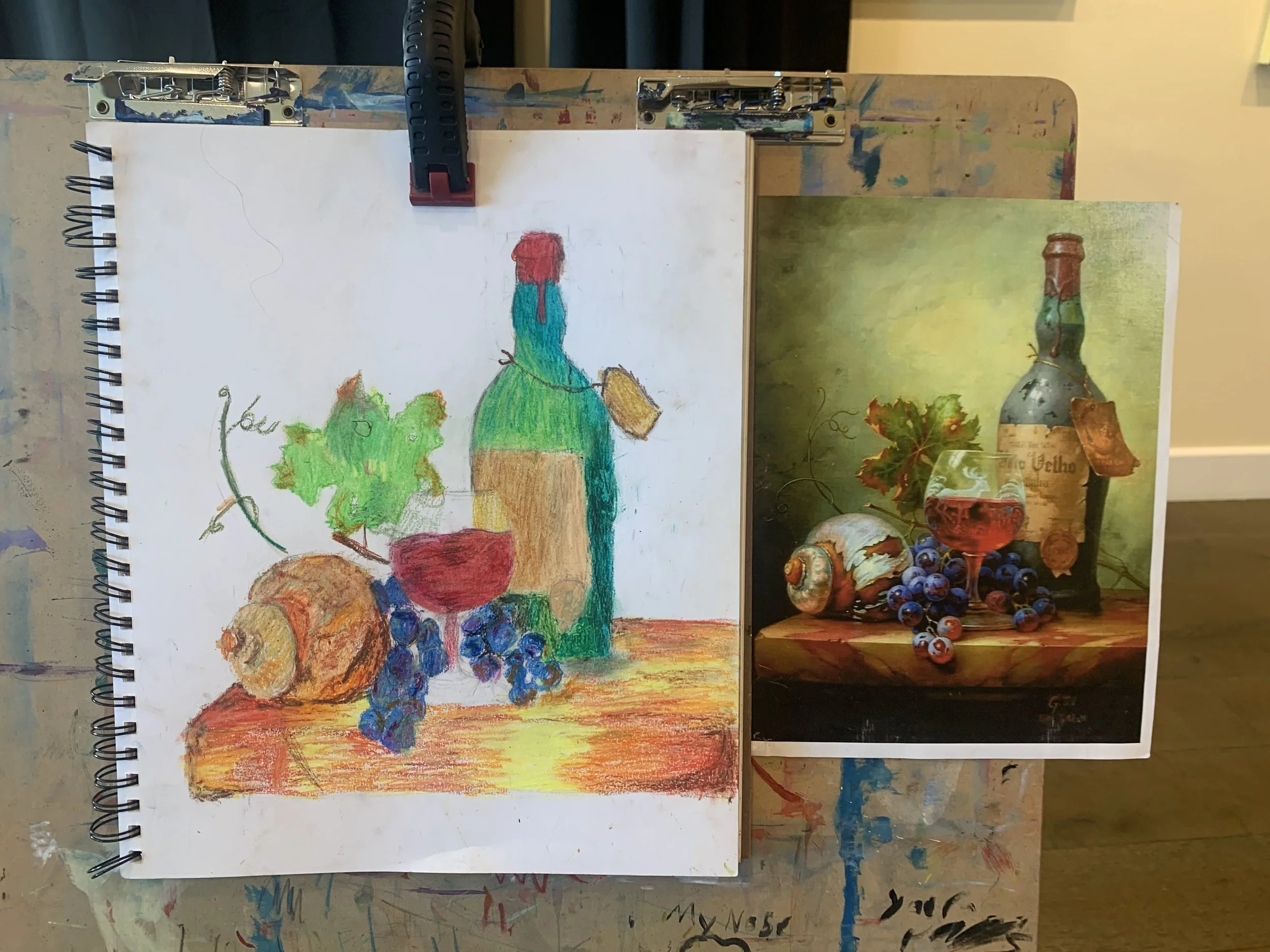 Drawing in progress of a still life with a wine bottle, a glass of red wine, a seashell, a bunch of grapes, and a leafy plant on a wooden surface, with a printed reference photo beside it.