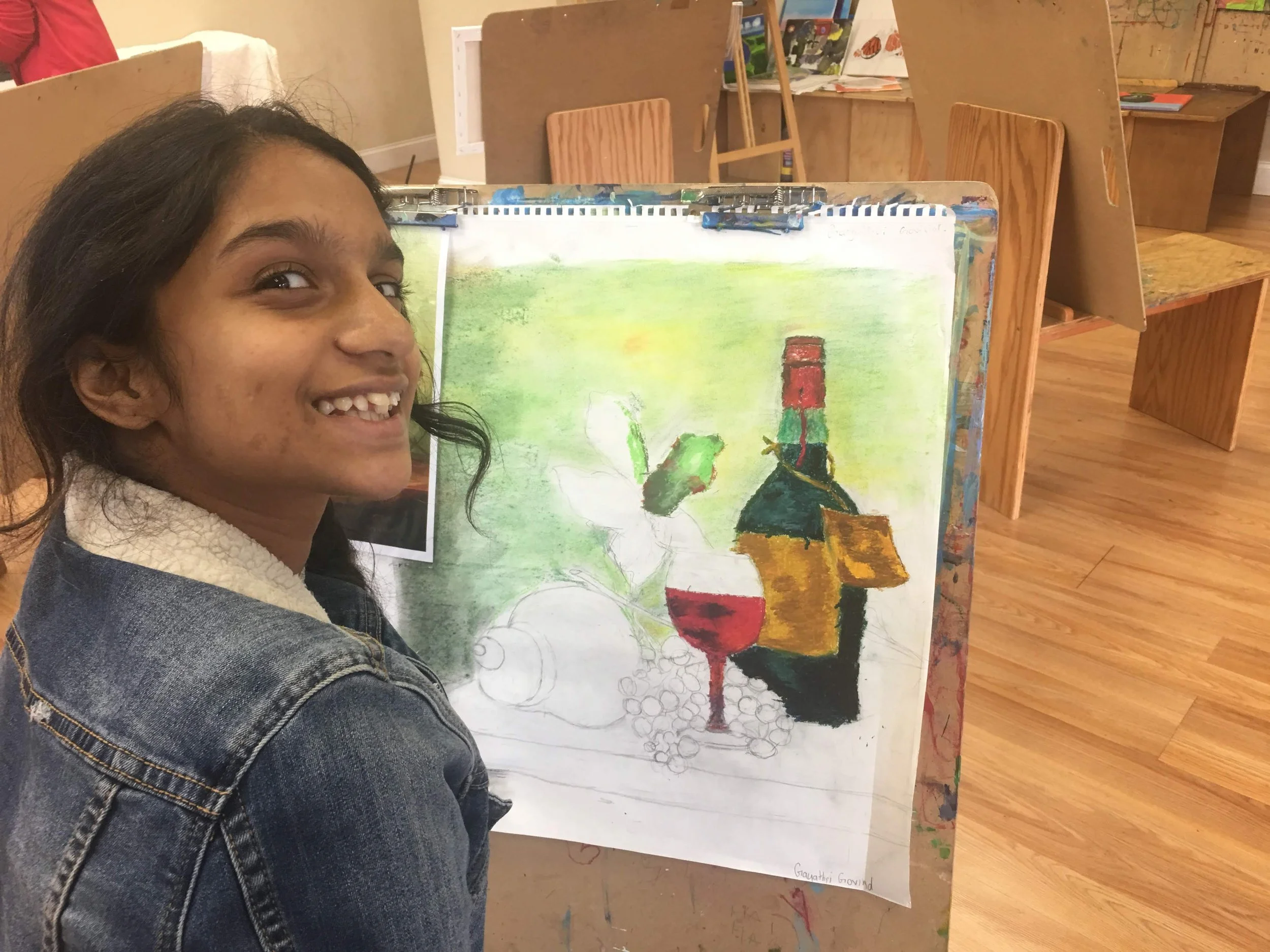 A young girl with dark hair, wearing a denim jacket with a fleece collar, smiling and showing missing front teeth, sitting next to a drawing of a still life scene on an easel in an art classroom. The drawing features a wine glass, grapes, a green bot