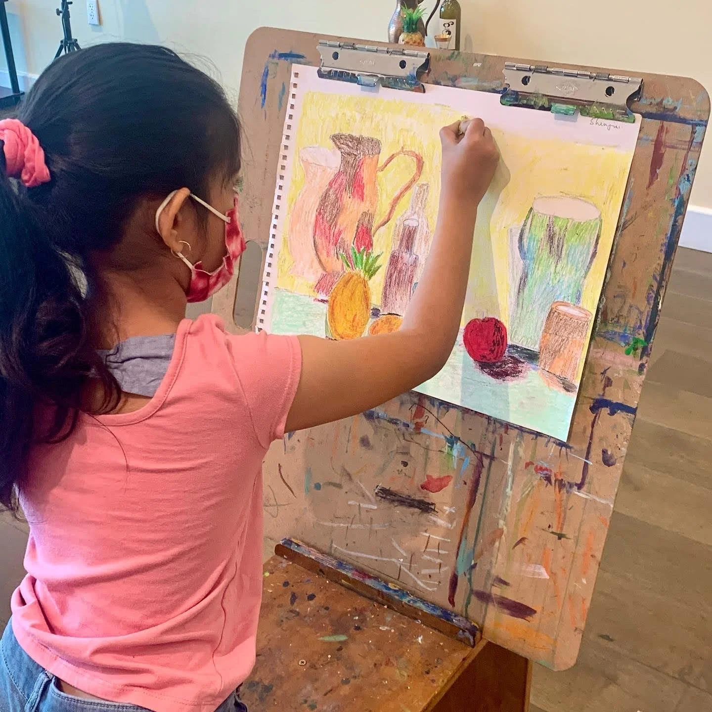 A young girl wearing a pink shirt, glasses, and a pink face mask is drawing with pastels on a large piece of paper attached to an easel. She is creating a colorful still life scene with a pitcher, a pineapple, an apple, and other objects.
