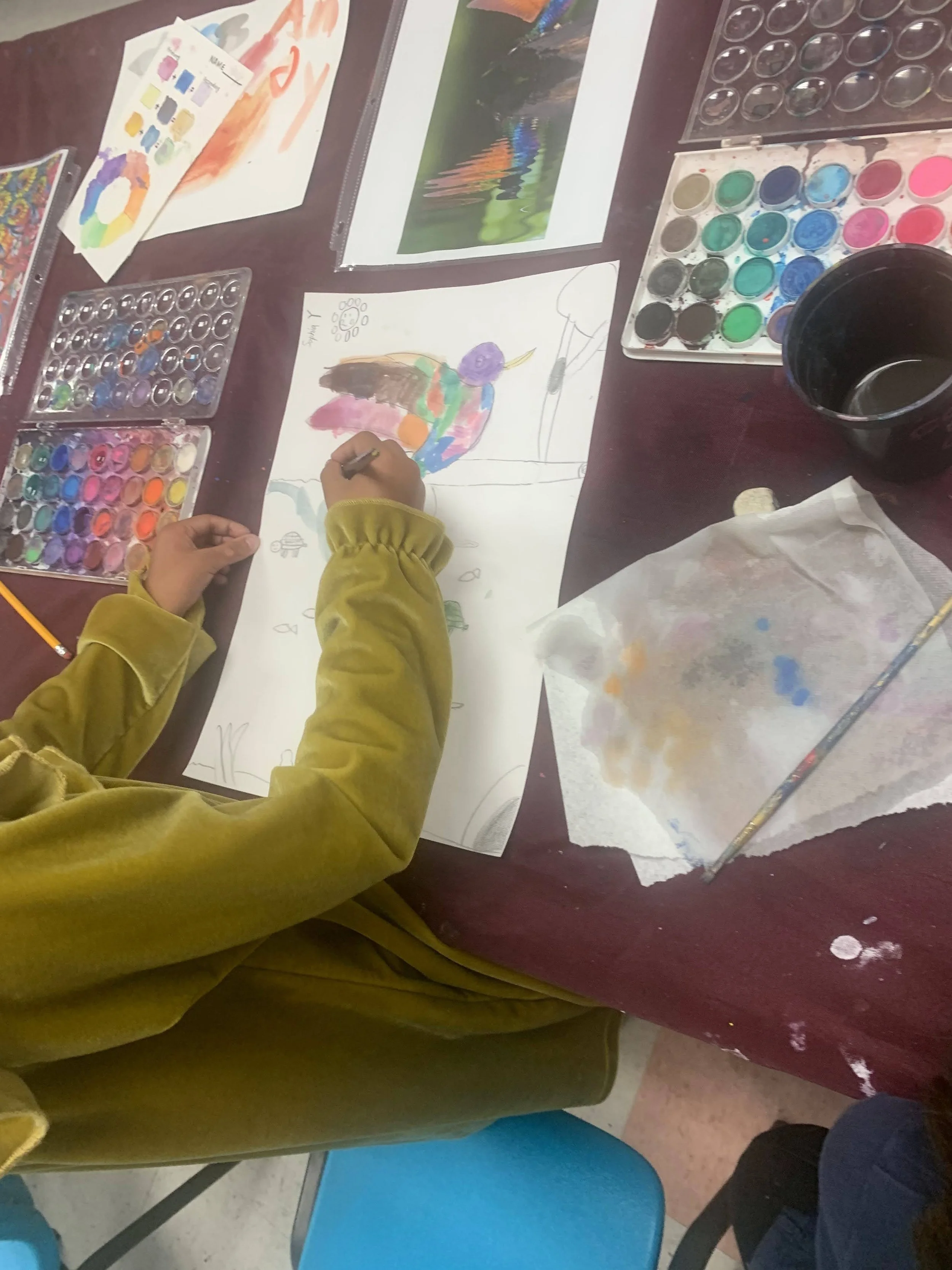 Child painting a colorful bird on a large sheet of paper surrounded by watercolor paints, brushes, and water containers at an art table.