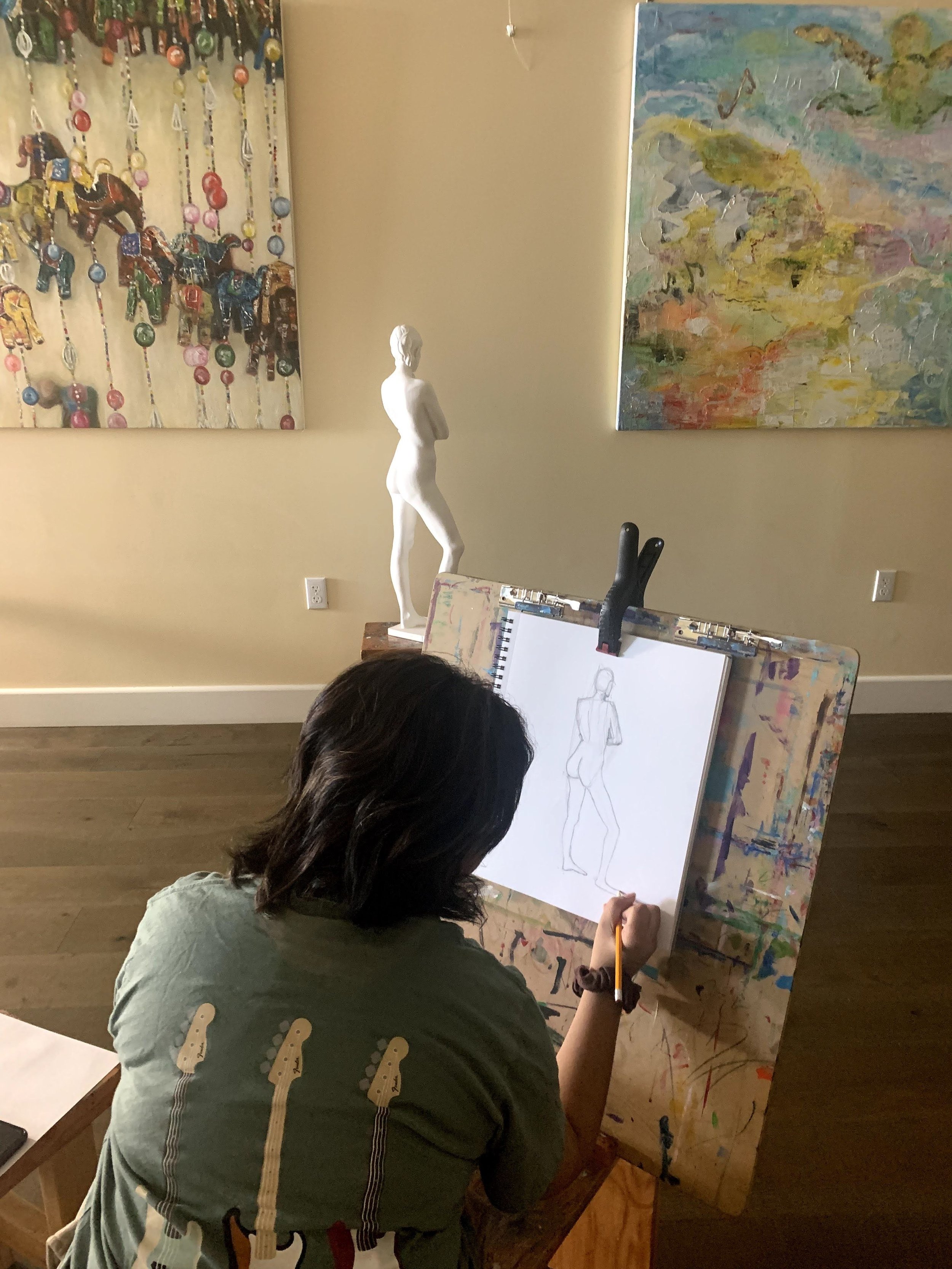 An artist sketches a human figure drawing on a sketchpad in an art studio, surrounded by abstract paintings and sculptures.