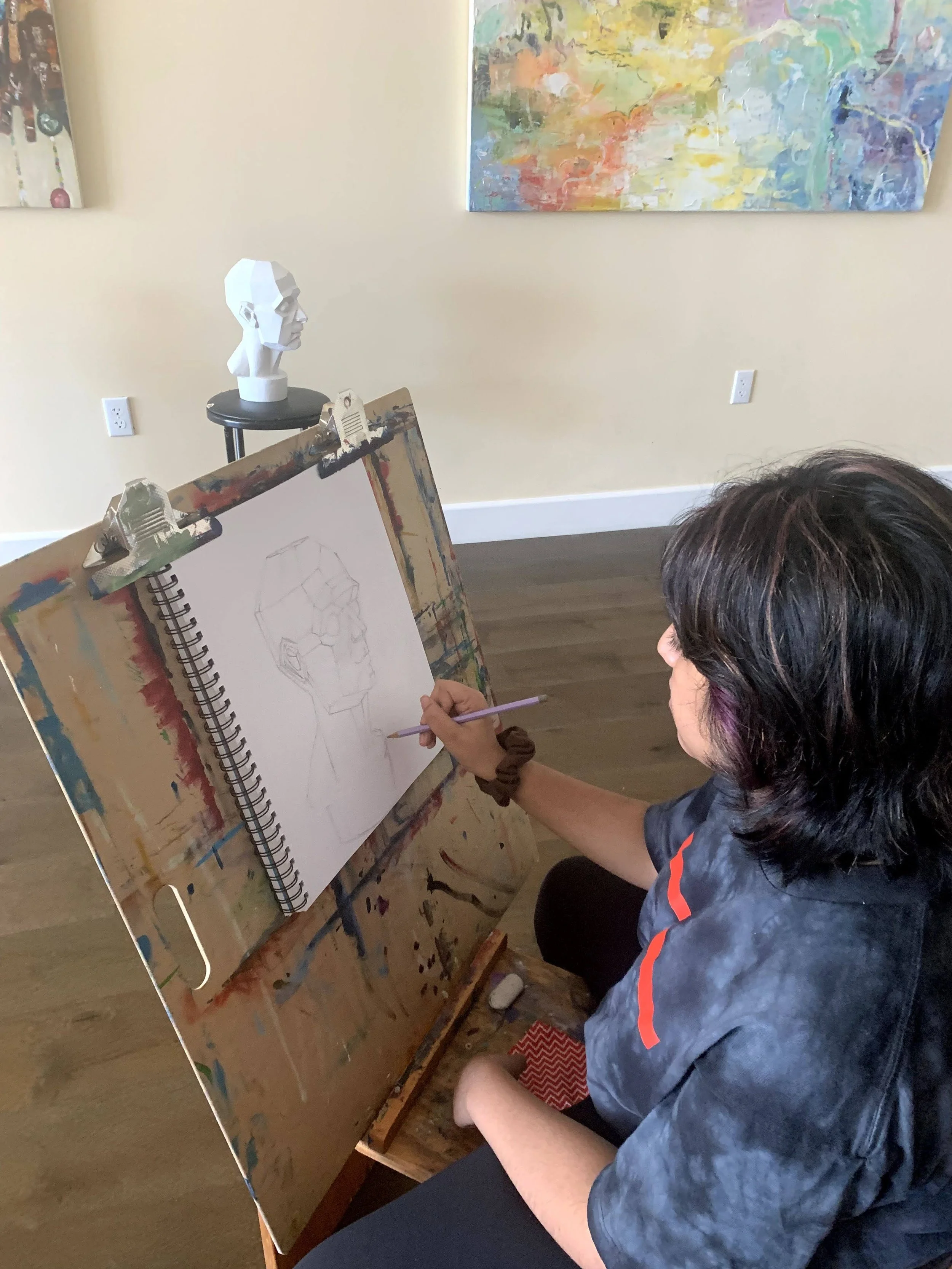 An artist sketches a portrait on a spiral sketchpad set on a palette-stained easel in a room with abstract artwork on the wall. A white bust sculpture is placed on a small black table behind the artist.