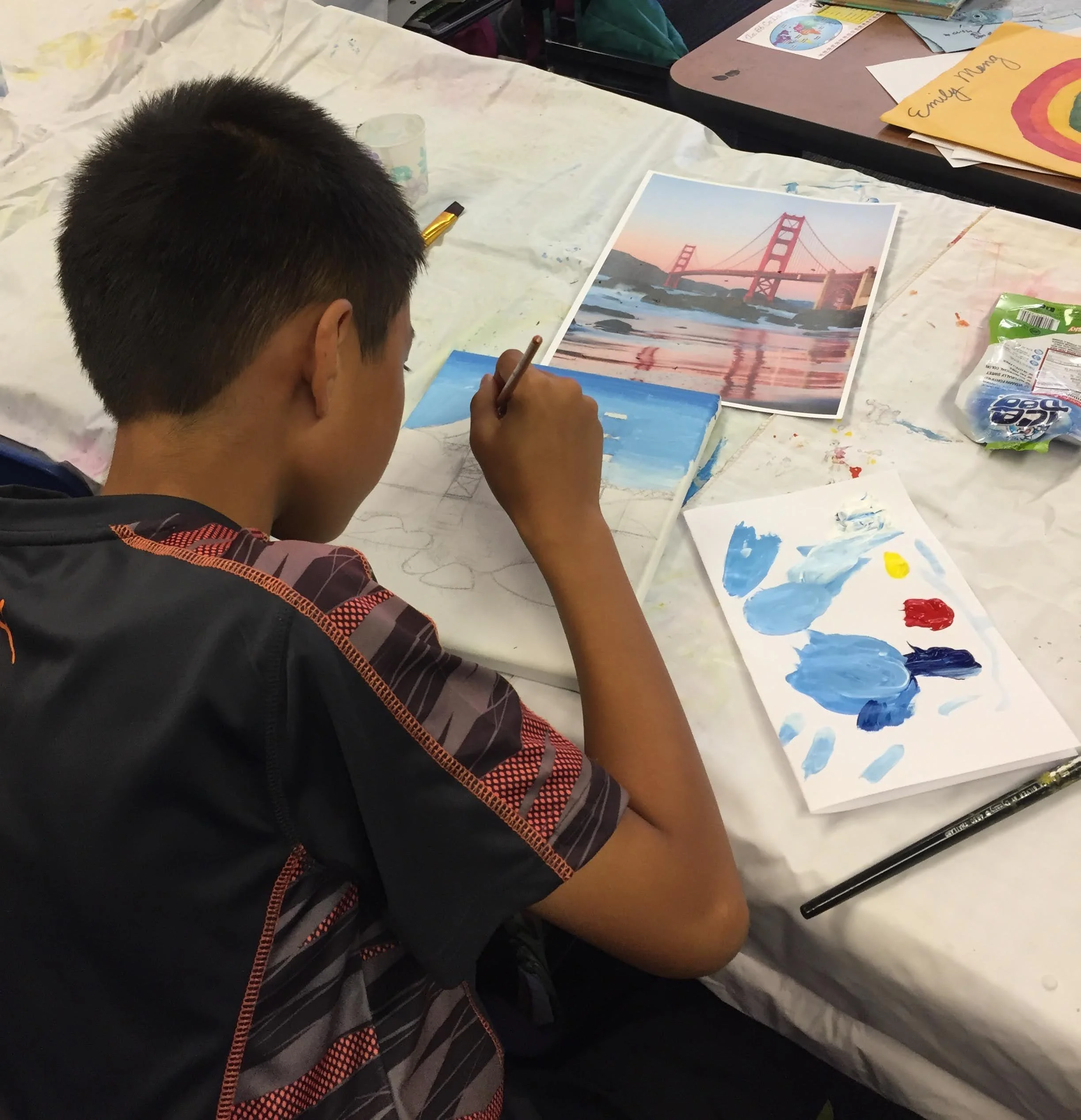 A boy painting at a table with a reference photo of the Golden Gate Bridge in front of him.