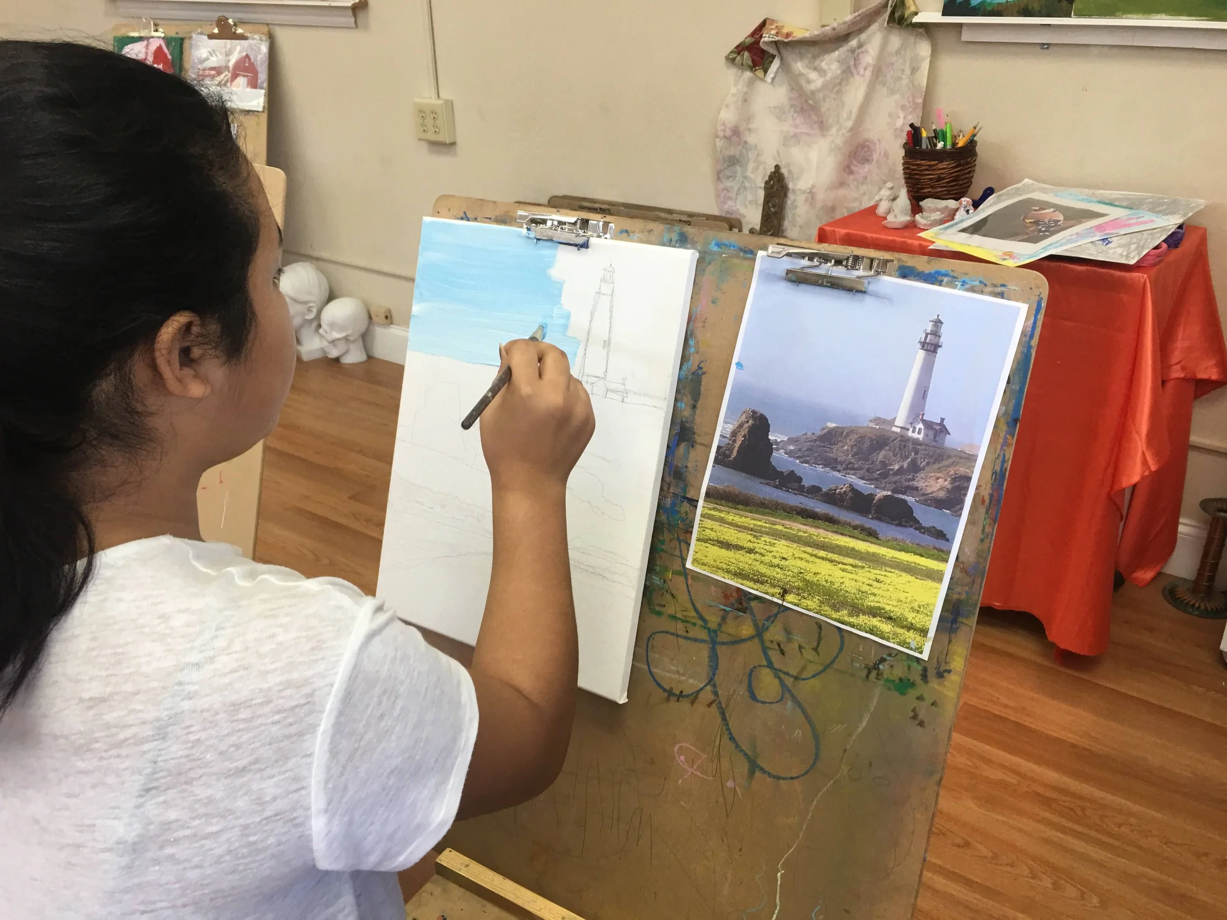 A woman is painting a lighthouse scene on a canvas, with a reference photo of a lighthouse on display beside it. She is seated at an easel in a cozy art studio.