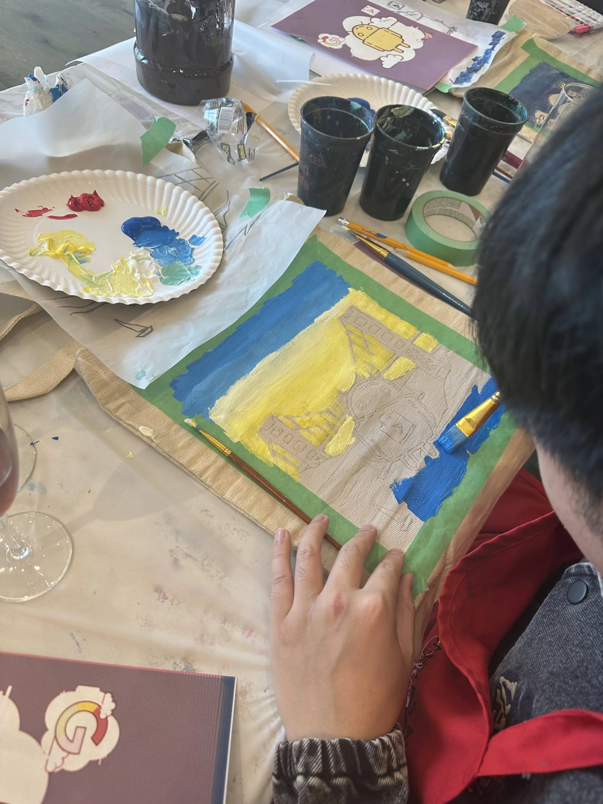 Person painting a landscape on a canvas with blue and yellow acrylic paint, surrounded by paint supplies, brushes, cups of black paint, tape, and paper on a table.