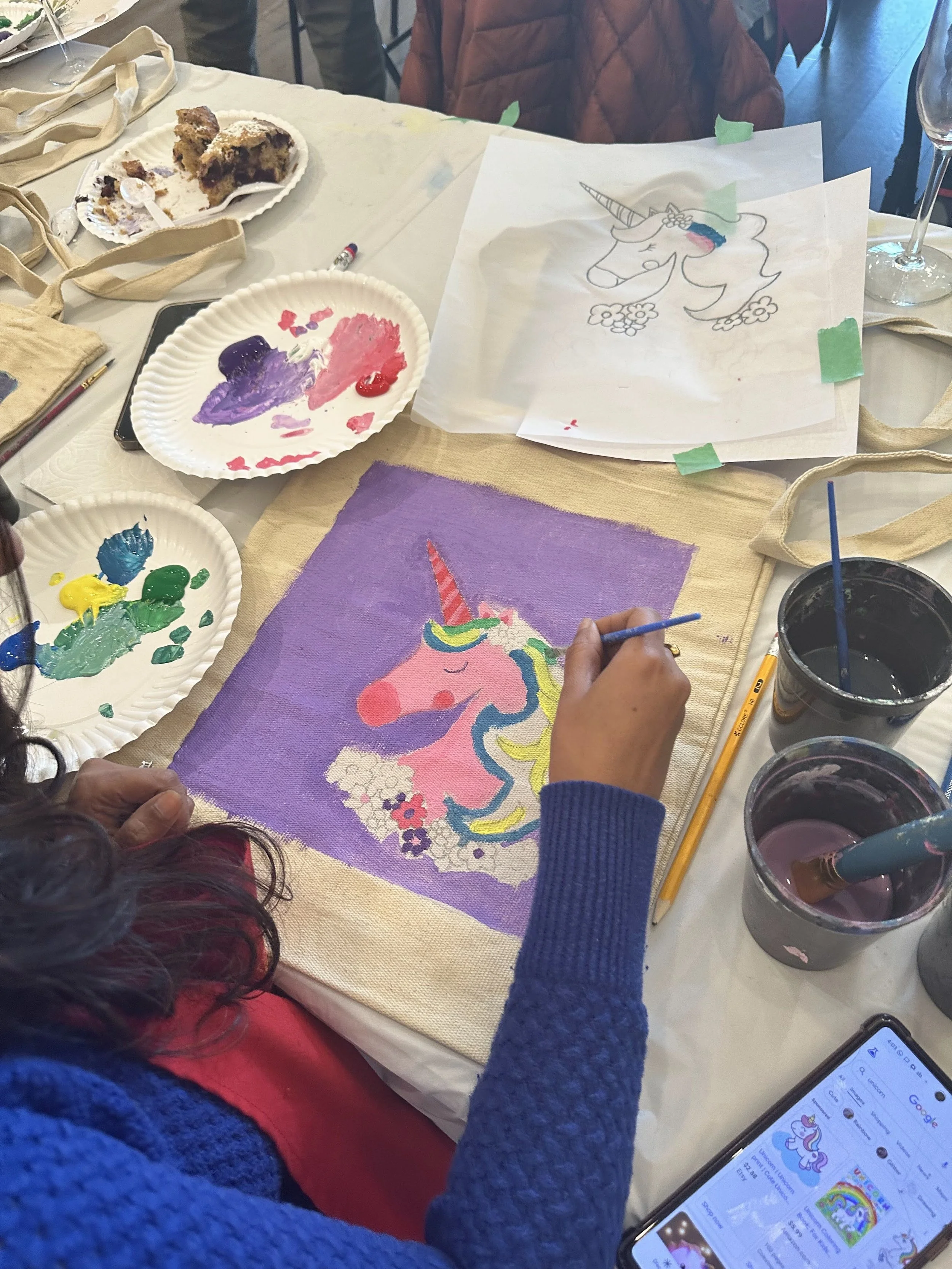 Person painting a colorful unicorn on a canvas, with painting supplies and a printed unicorn sketch on the table