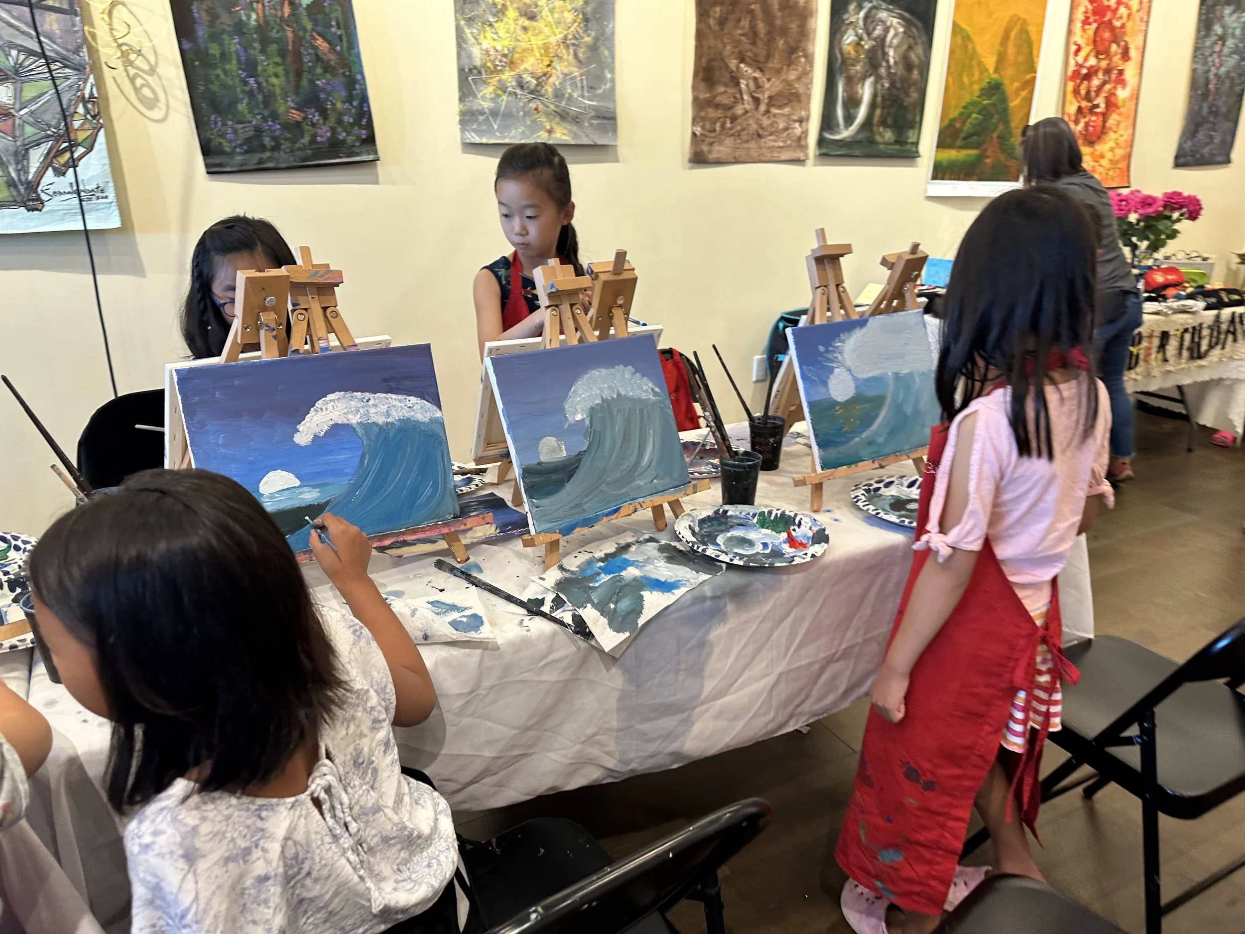 Children painting ocean waves on small canvases at a painting class, with colorful artwork hanging on the wall behind them.