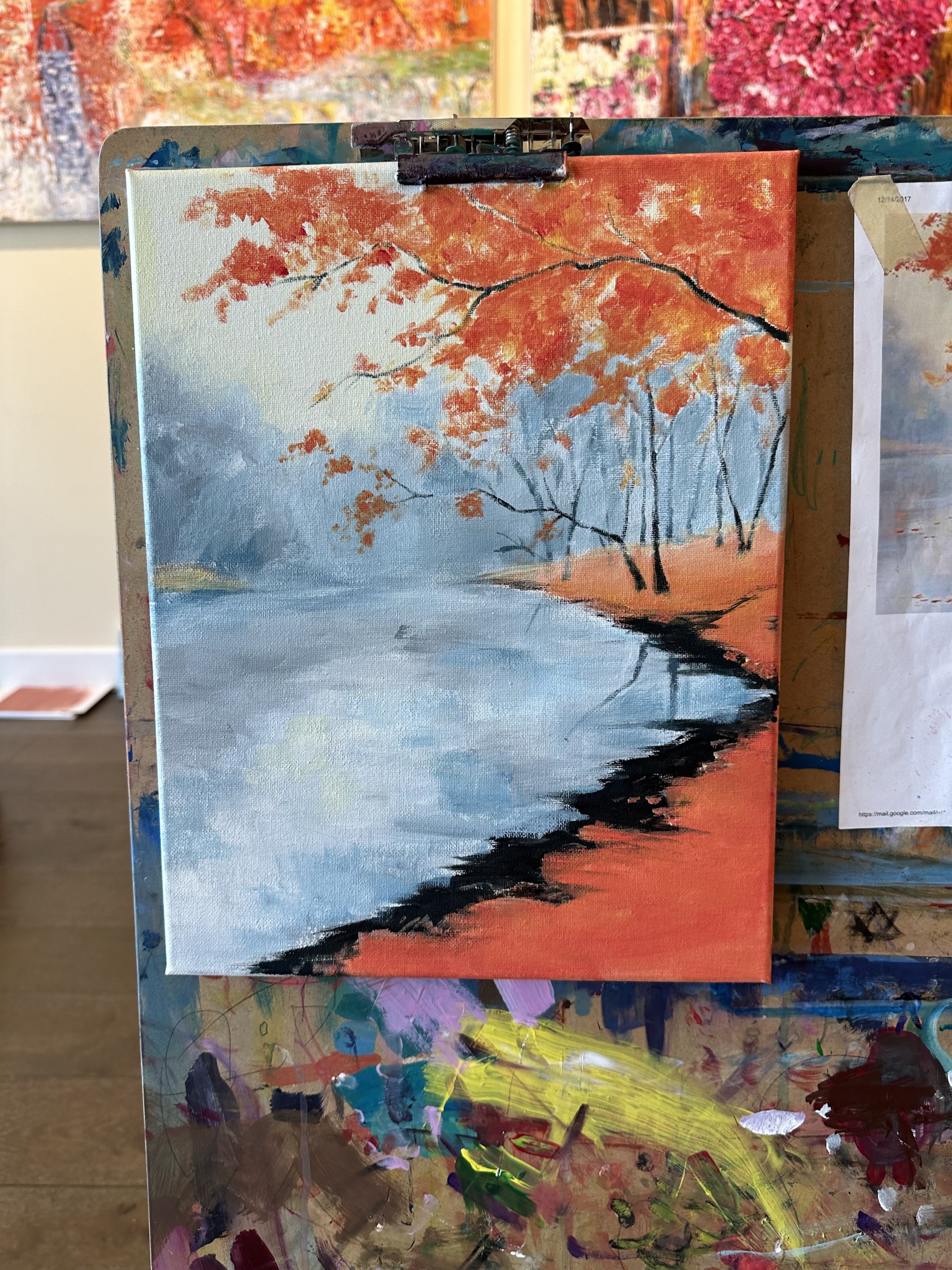 A landscape painting featuring a river with trees on the bank, bright orange and red autumn leaves, and a partly cloudy sky.