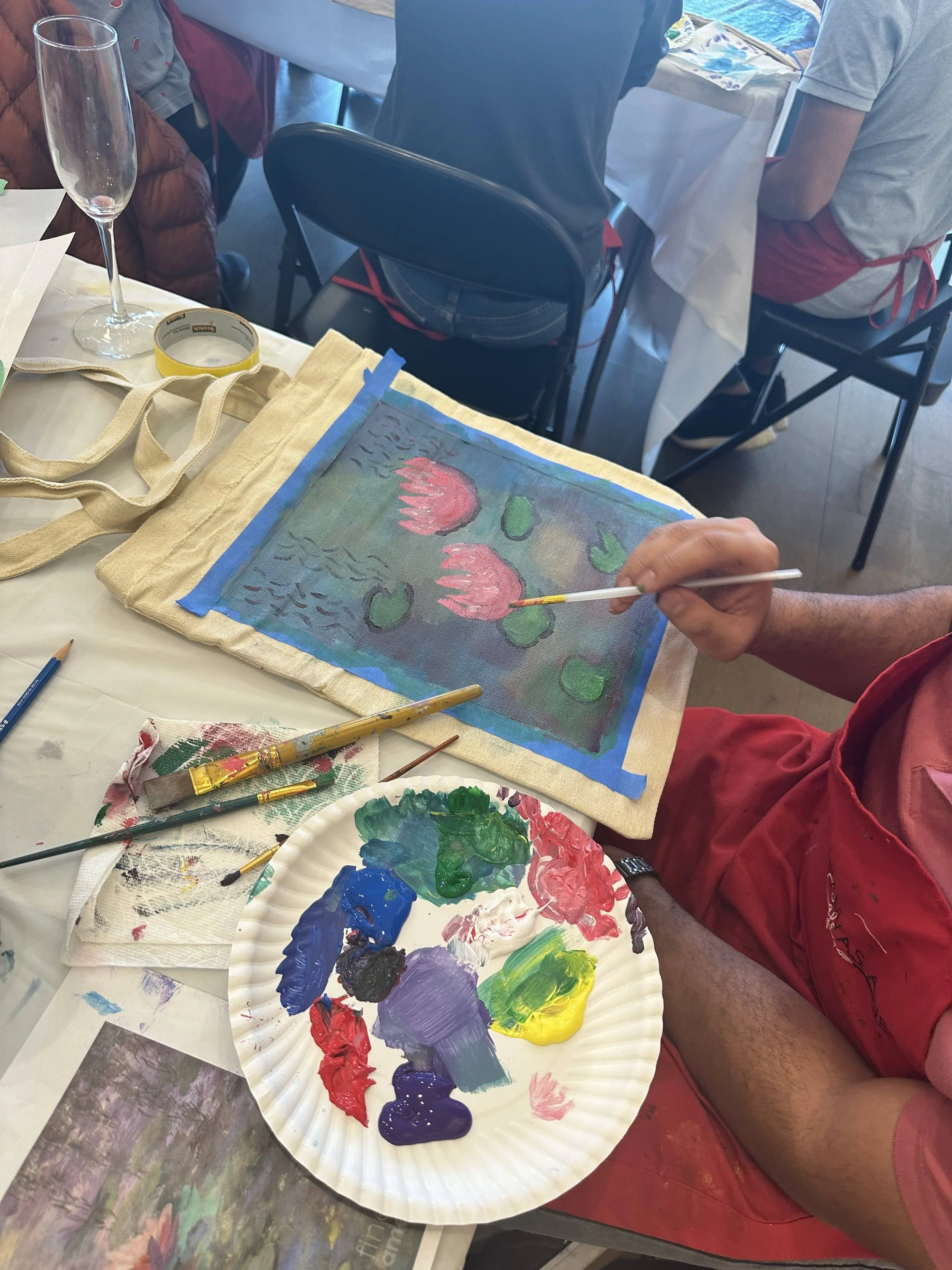 Person painting water lilies on fabric, using a brush with a palette of various colors nearby, at a table with other people.