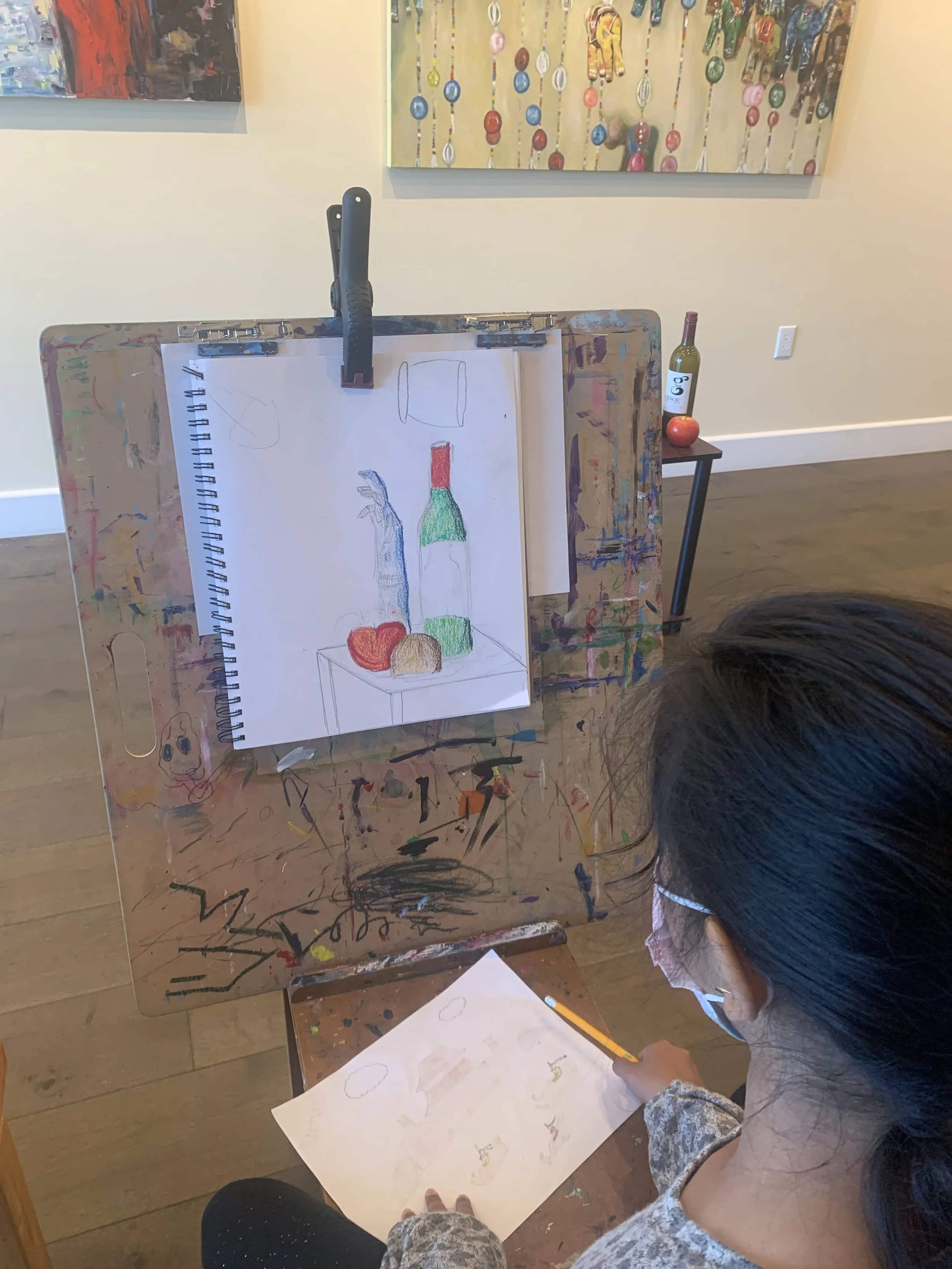 A young girl drawing on a sheet of paper while sitting in front of an artist's easel with a colorful still life drawing of a bottle, apple, and other objects. The easel is covered in paint splatters and stains. In the background, a table with a bottl