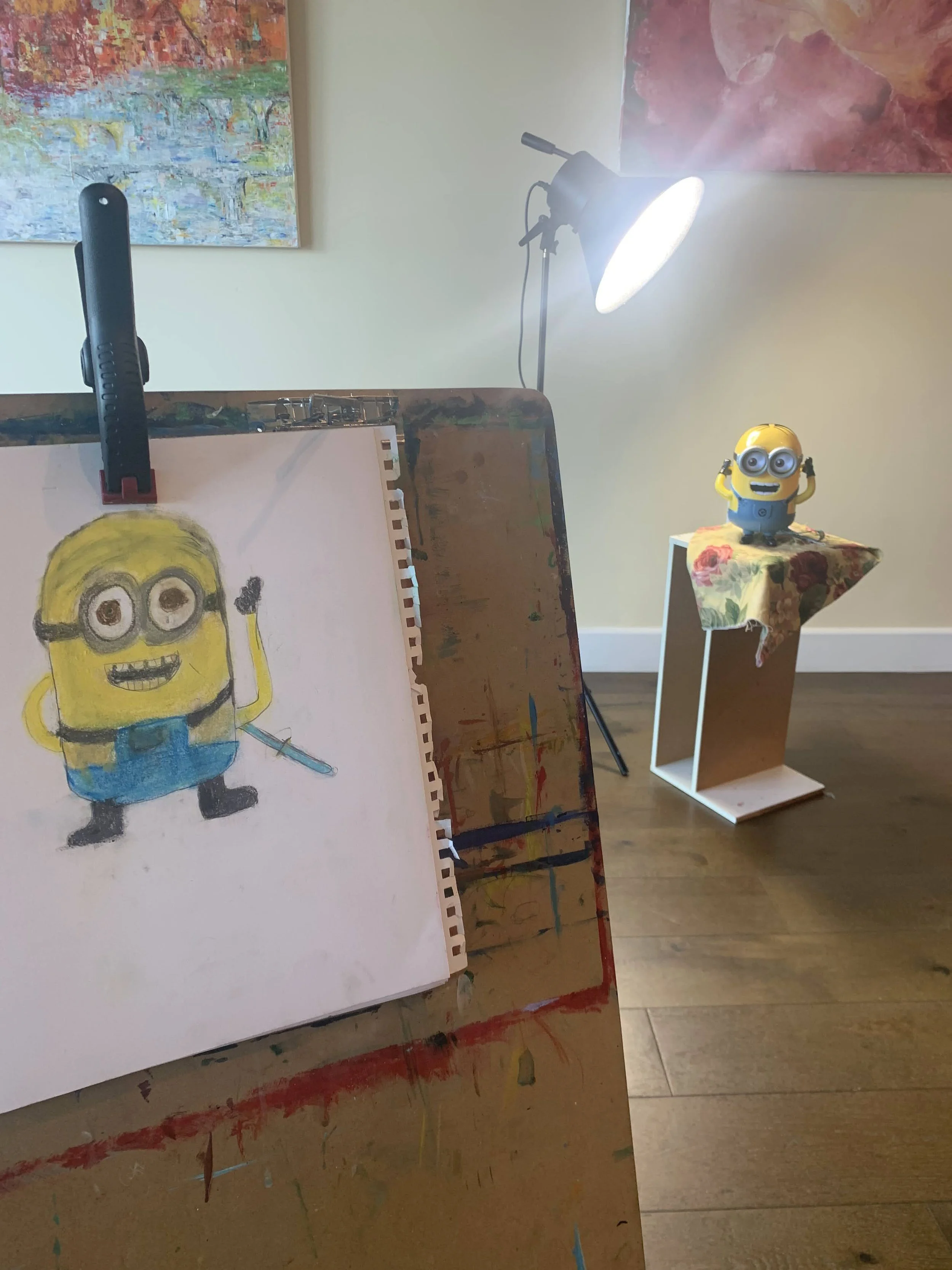 A workspace with a drawing of a Minion character in progress, a Minion figurine on a small table, and a bright studio light in the background.