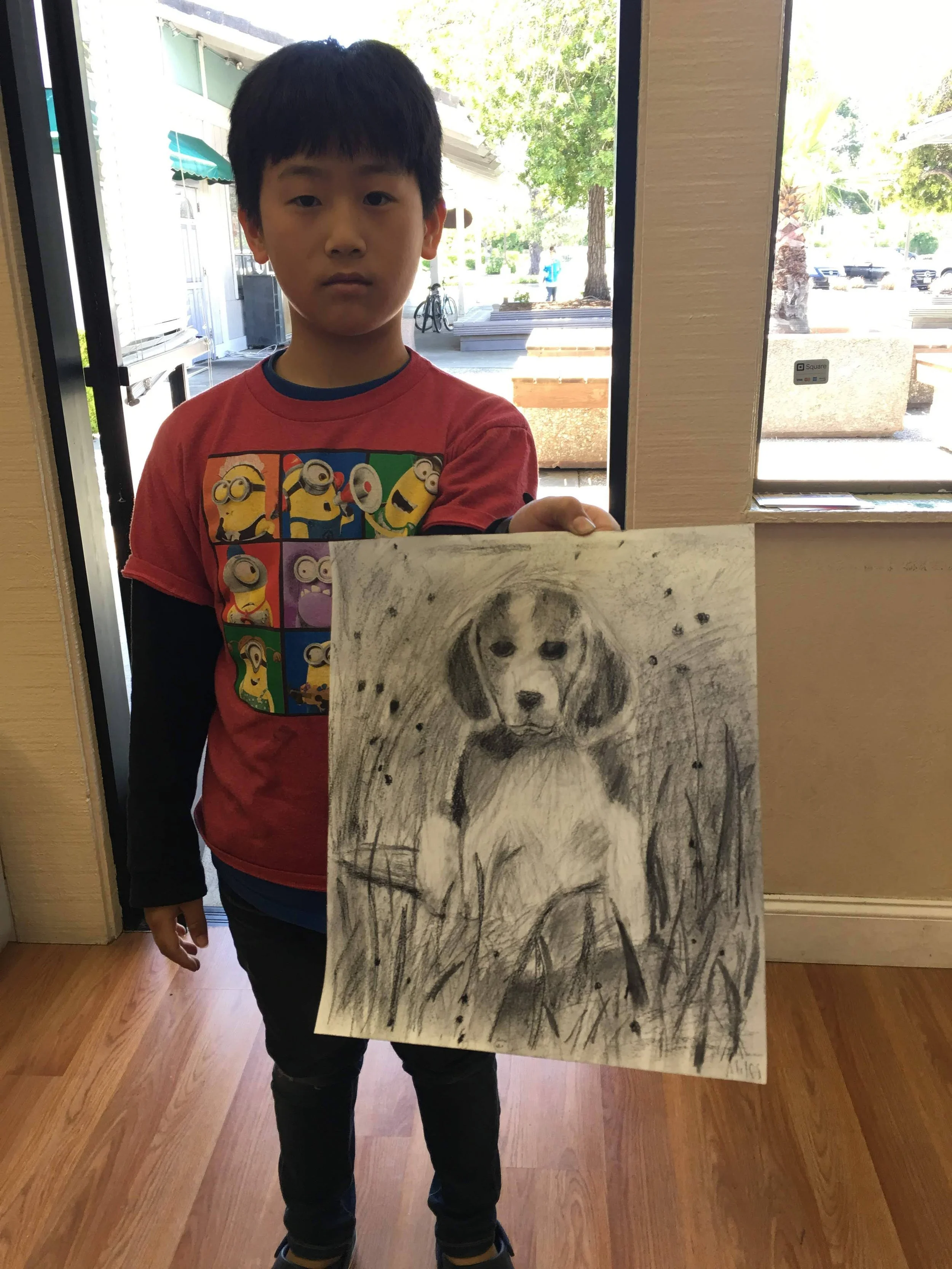 A young boy with dark hair stands inside a building, holding a black and white charcoal drawing of a dog sitting in tall grass. The boy is wearing a red shirt with Minions characters on it, and the background shows a door, window, and an outdoor scen