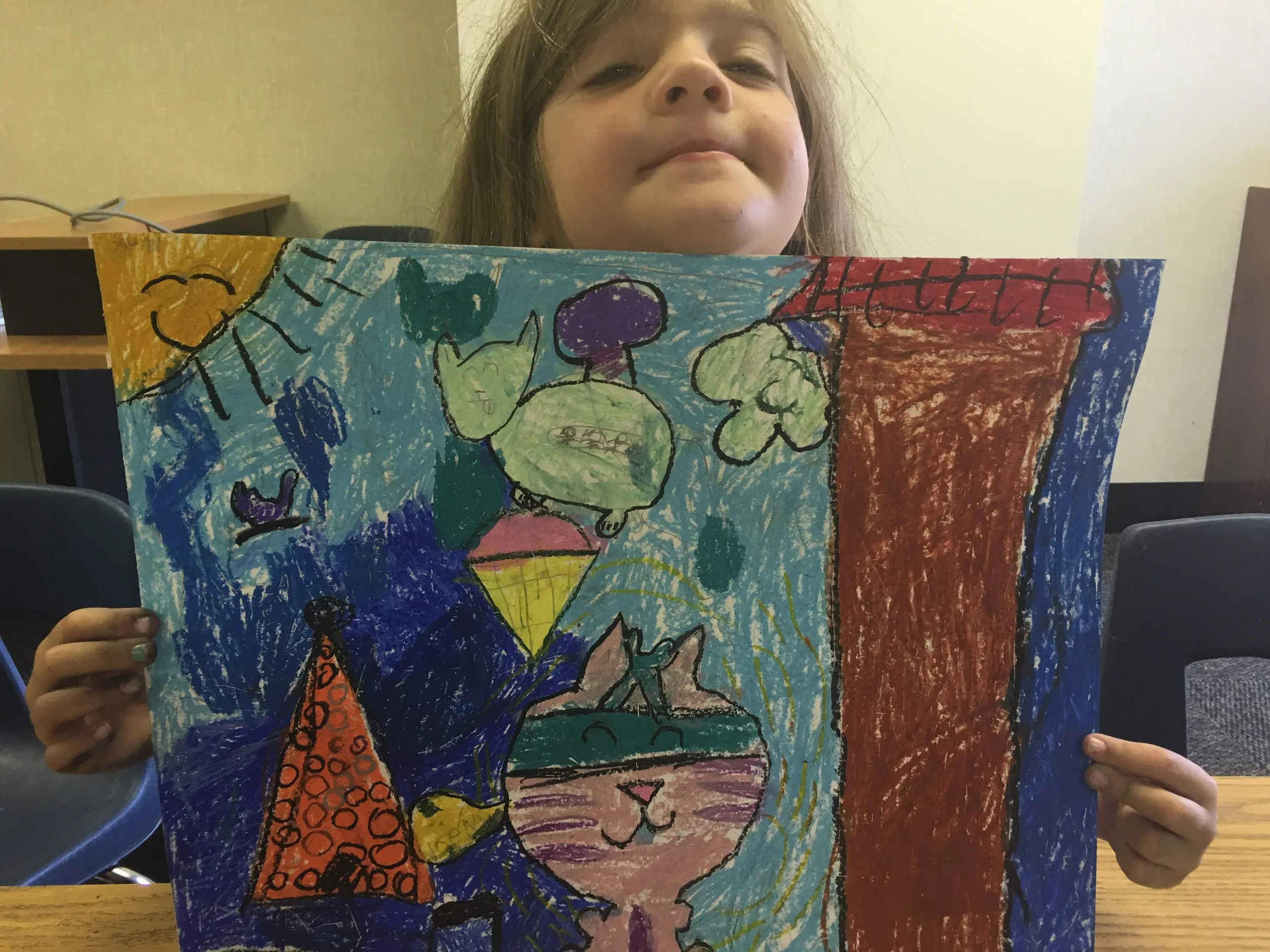 Child holding a colorful drawing featuring cats, party hats, the sun, clouds, and a large brown object, all sketched with crayons.