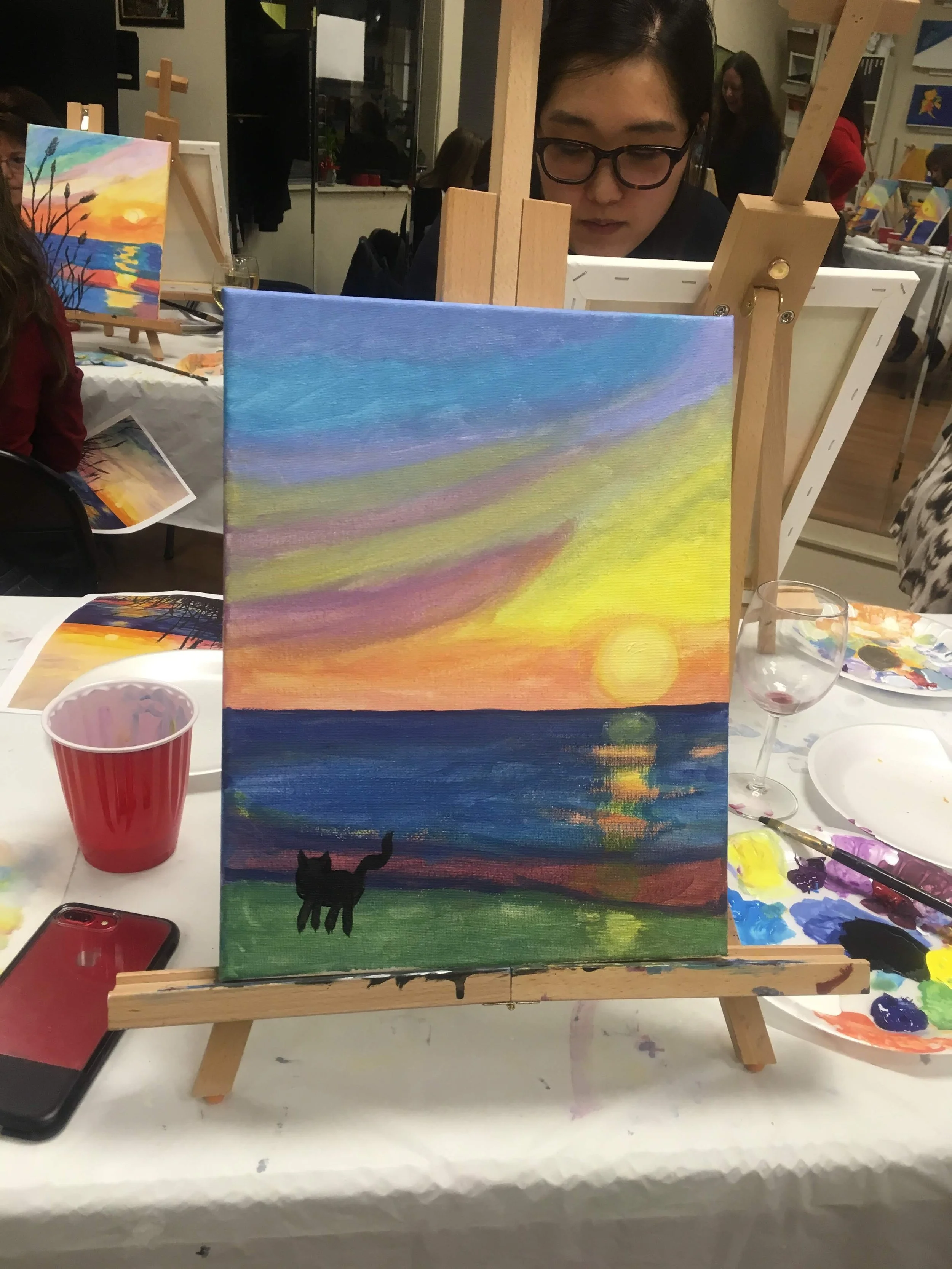 Painting of a sunset over water with a black cat silhouette in the foreground, on an easel at an art class.