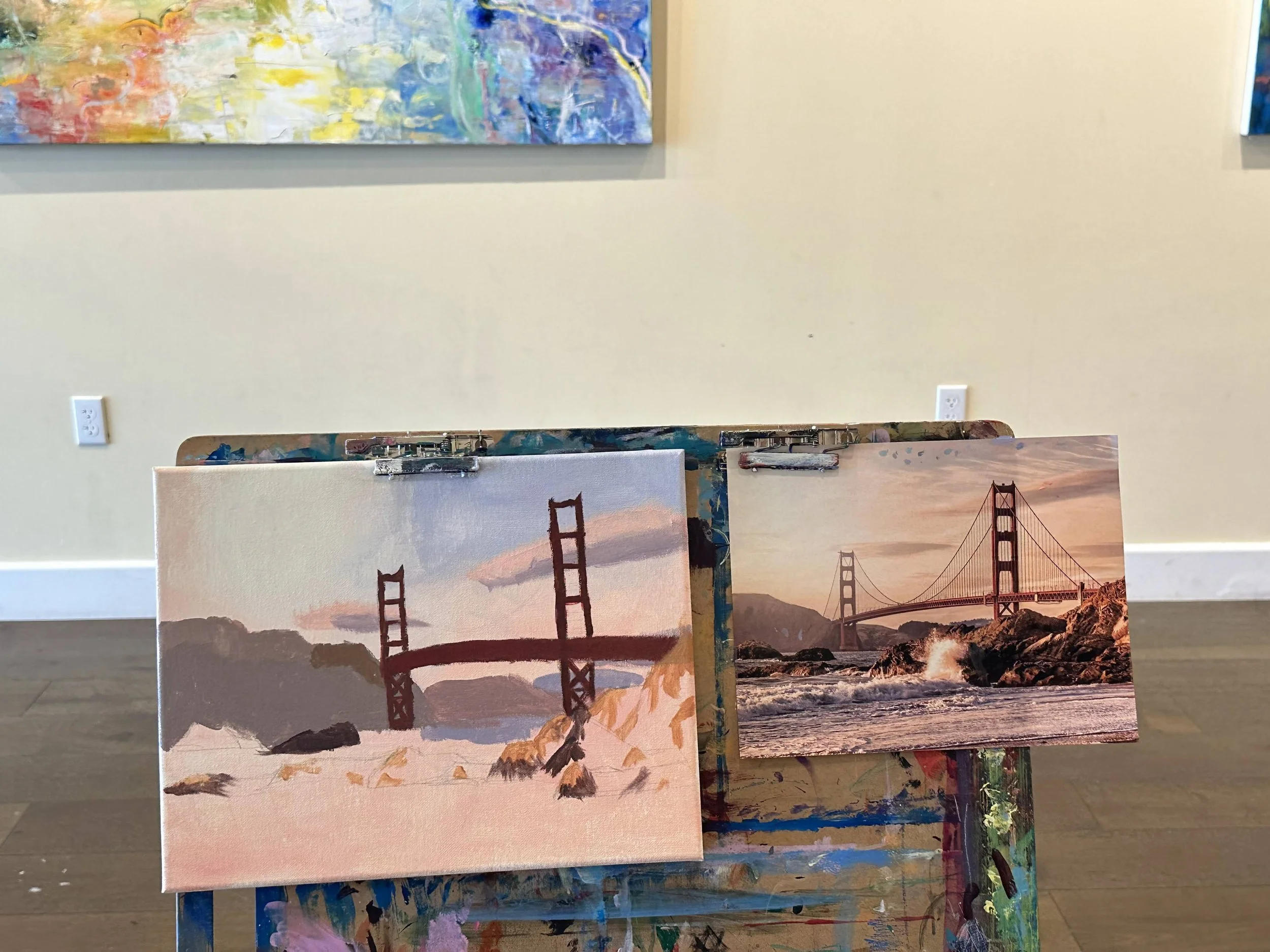 Two paintings of the Golden Gate Bridge, one in a realistic style and one in a watercolor style, are displayed on an artist's easel in an art gallery.