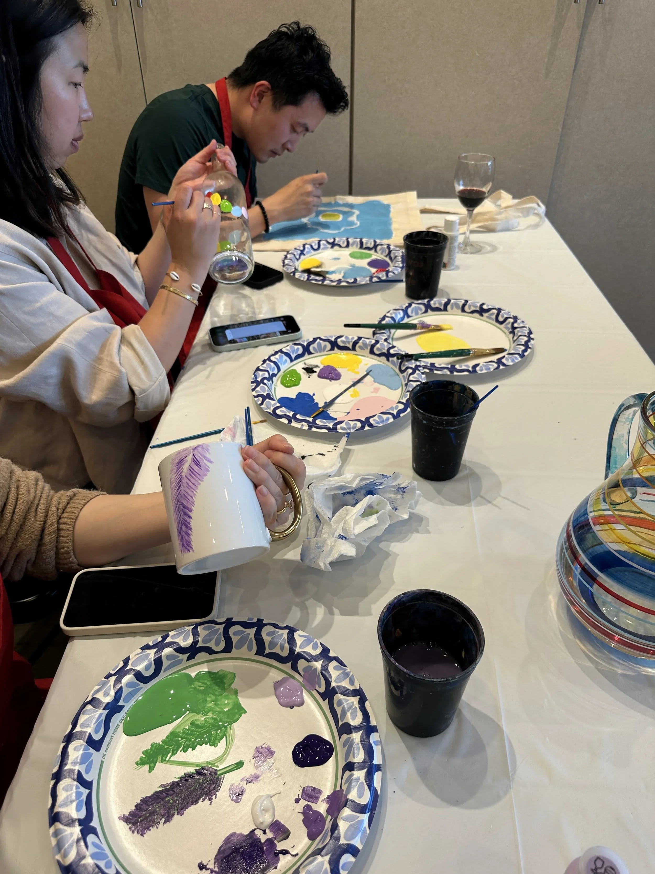 People sitting at a table painting ceramic mugs and plates with paints and brushes, with paintings and painting supplies visible.