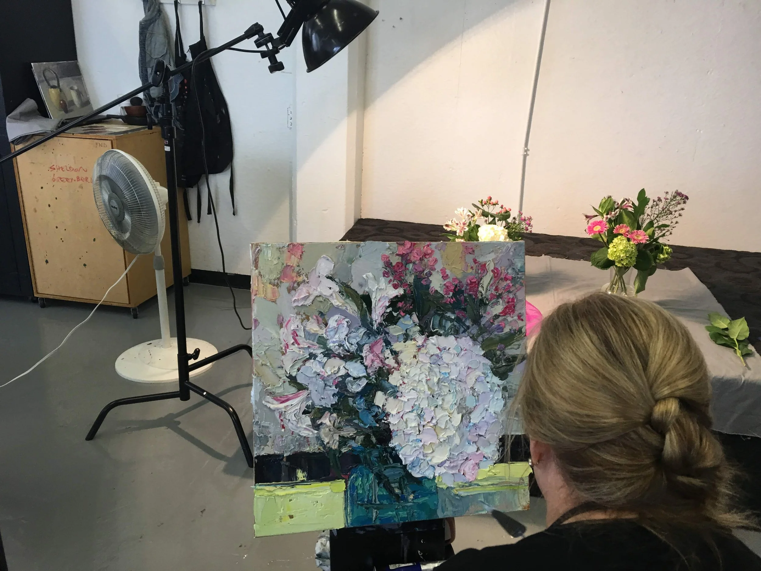 Person painting a floral still life with white and pink flowers on canvas. Art supplies and floral arrangements are on a table in the background.