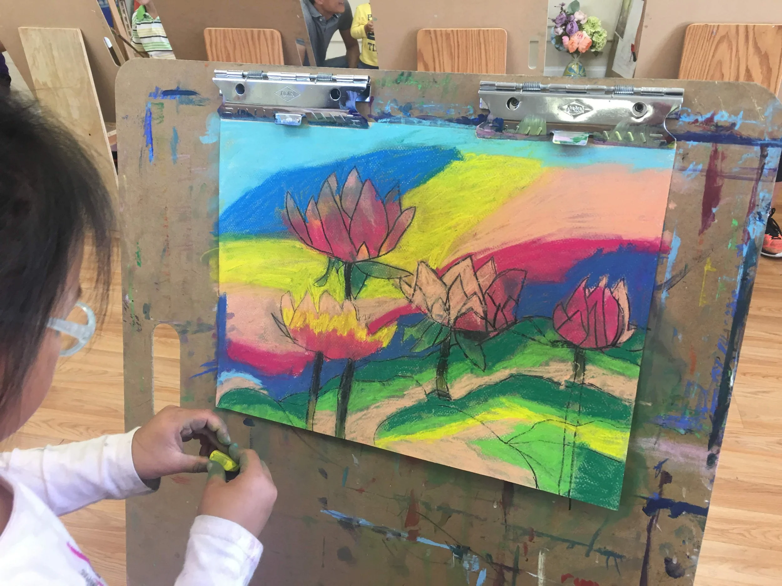 Child working on a colorful painting of lotus flowers on a board, using chalk or pastel, with people and chairs visible in the background.