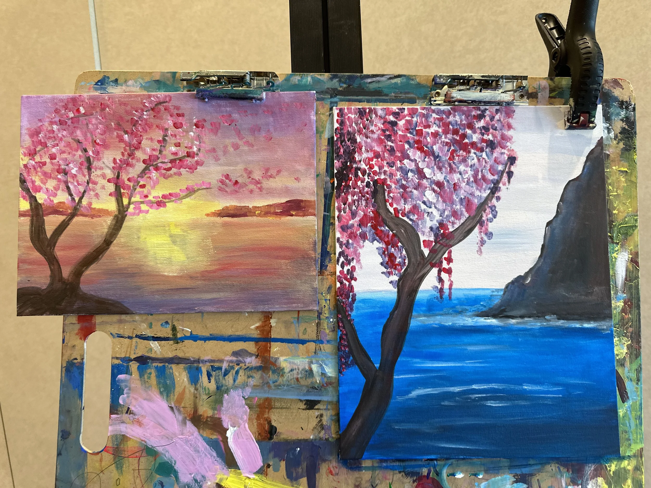 Two paintings of cherry blossom trees, one with pink blossoms at sunset over water, and the other with pink blossoms near a rocky coastline and blue sea.