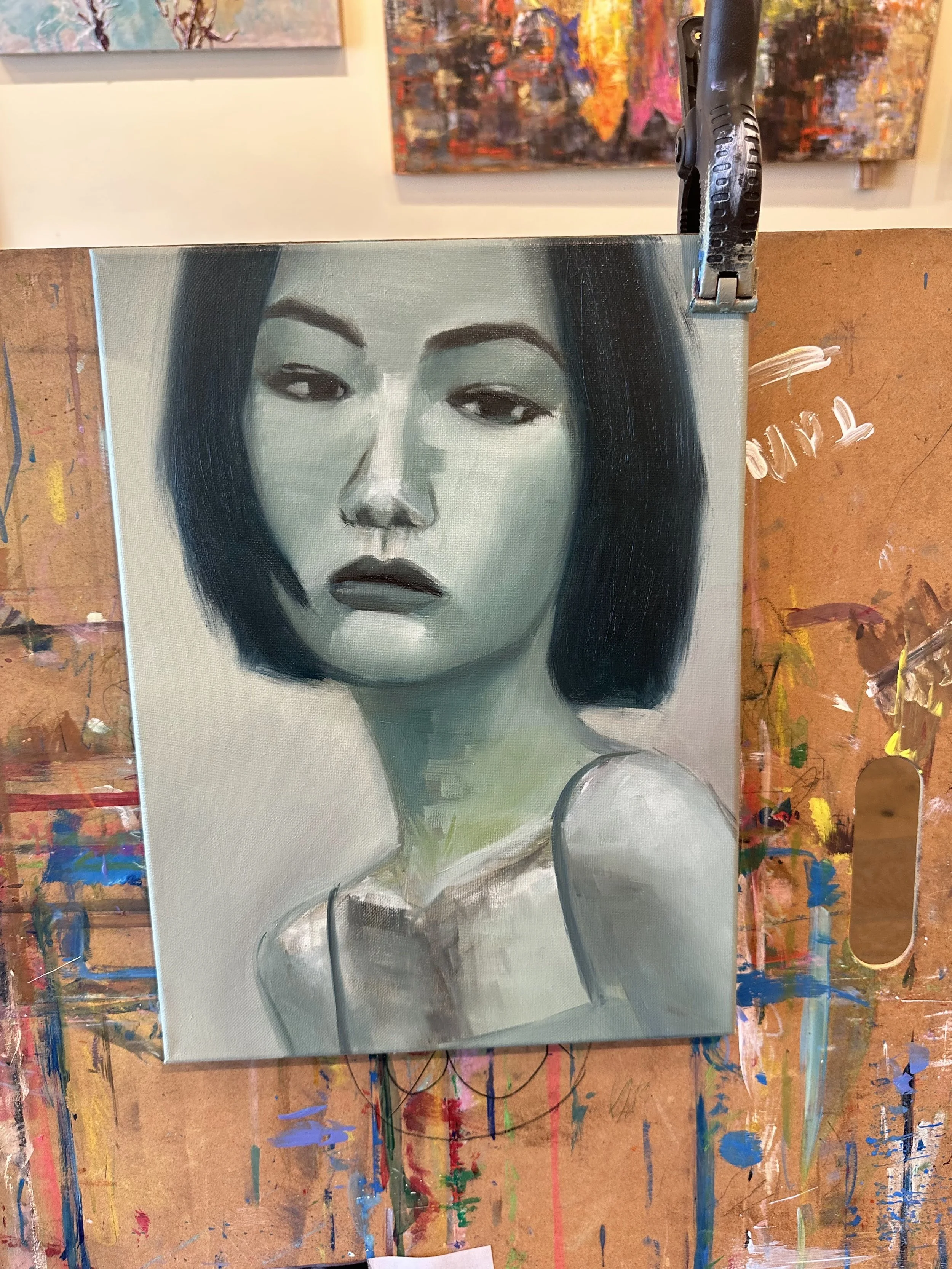 A grayscale portrait painting of a woman with short black hair, wearing a sleeveless top, displayed on an easel in an art studio.