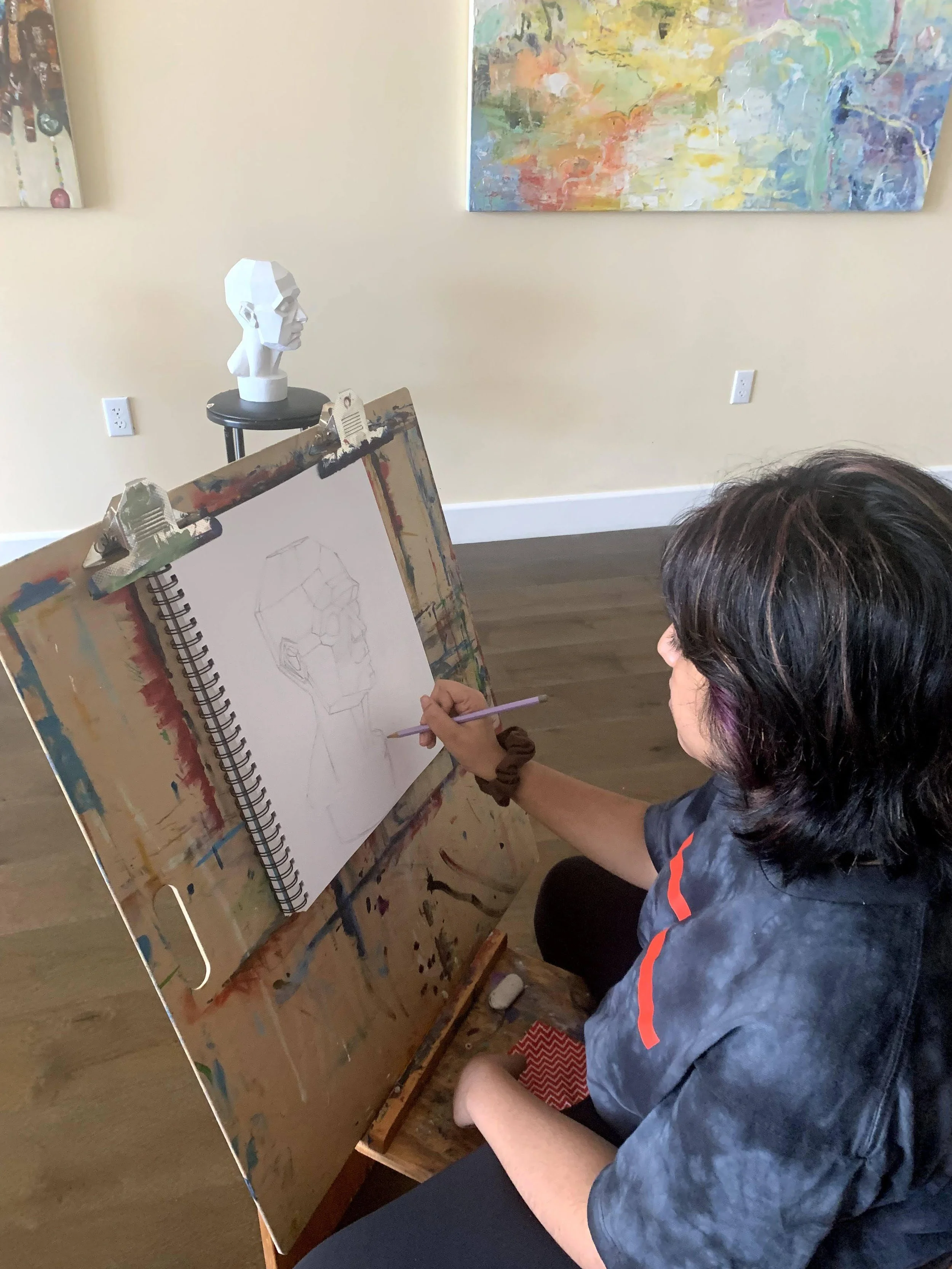 Person drawing a sketch of a head on a spiral notebook, with a pedestal displaying a geometric bust sculpture in the background, colorful abstract paintings on the wall, and a wooden floor.